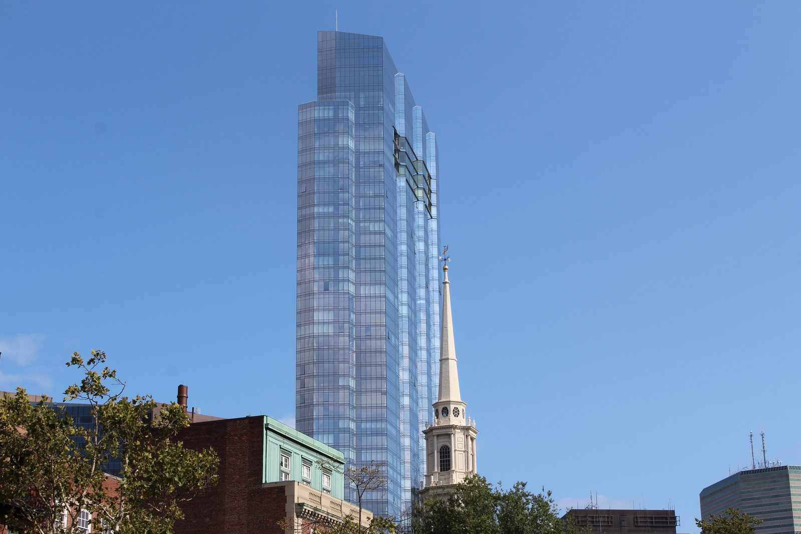 Millennium Tower's blue glass facade rising above a white church steeple and older brick buildings in downtown Boston