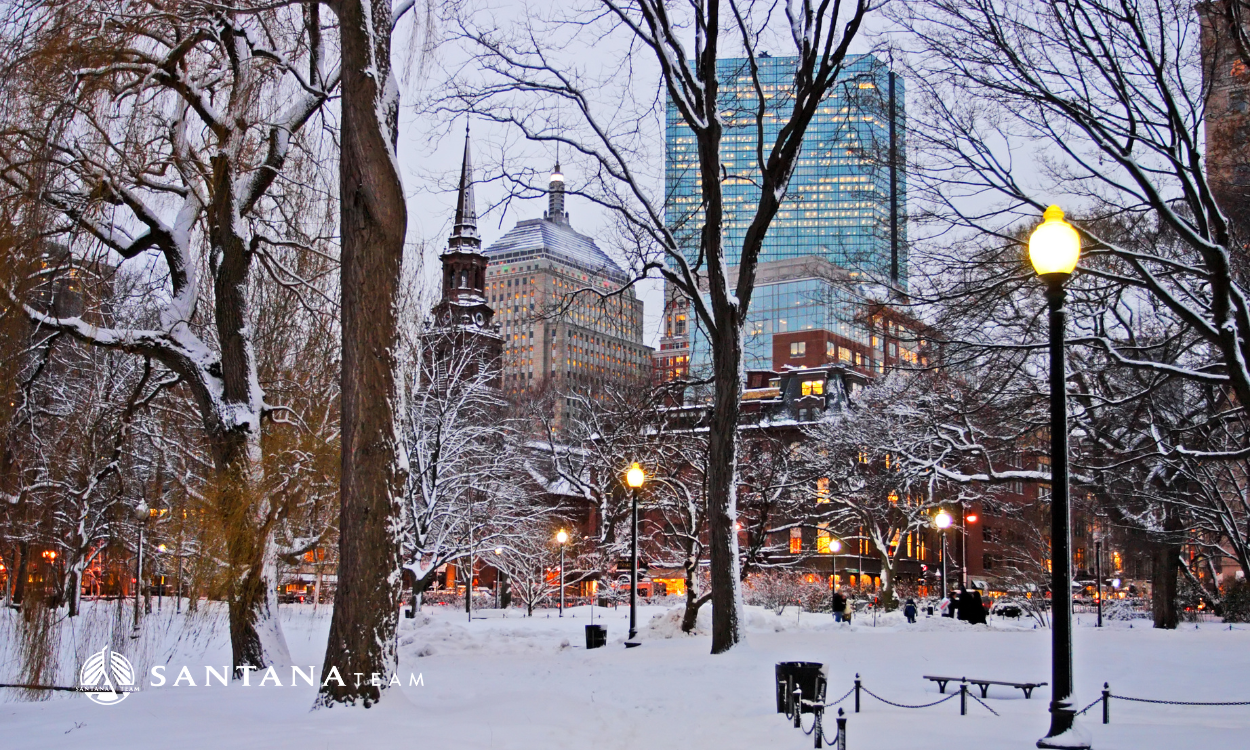 Boston Common winter scene with city skyline, snowy trees, and holiday lights at dusk
