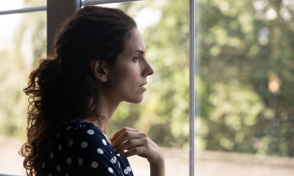 Person looking out a window thoughtfully, considering whether to make a move in the real estate market.