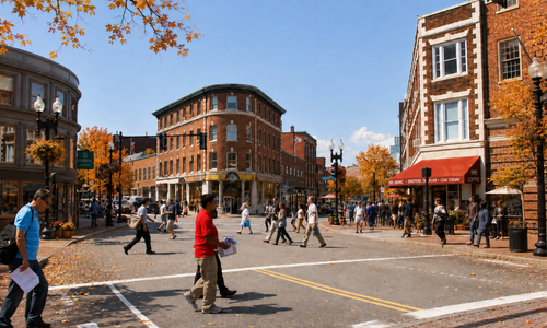 Autumn foliage and pedestrians in Harvard Square, Cambridge, with a few pumpkins and fall decorations along the streetscape.