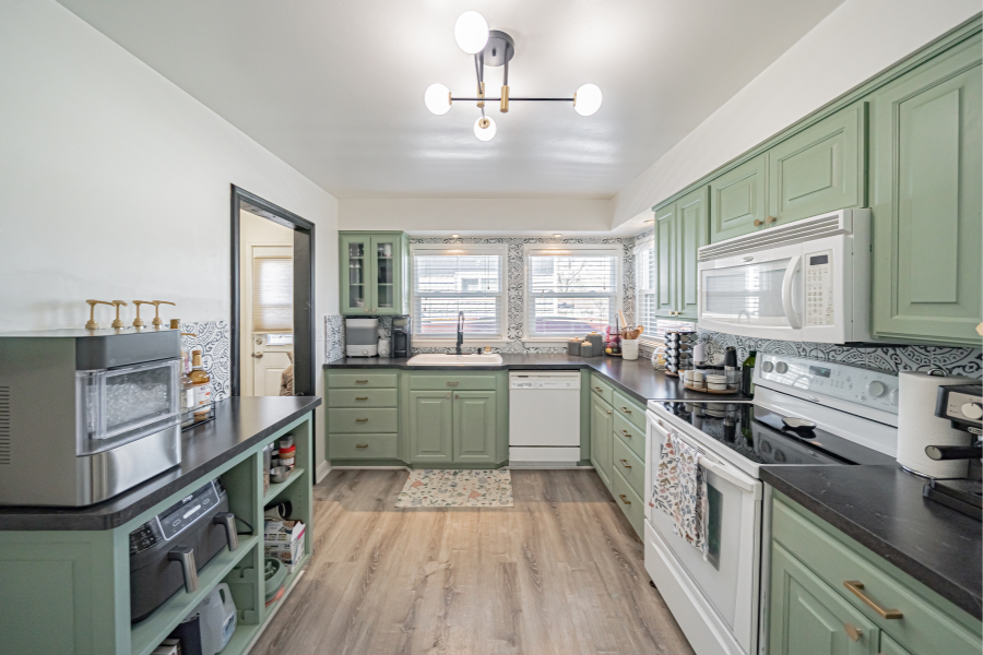 Bright kitchen with sage green cabinets, white appliances, black countertops, patterned tile backsplash, and three large windows providing natural light.