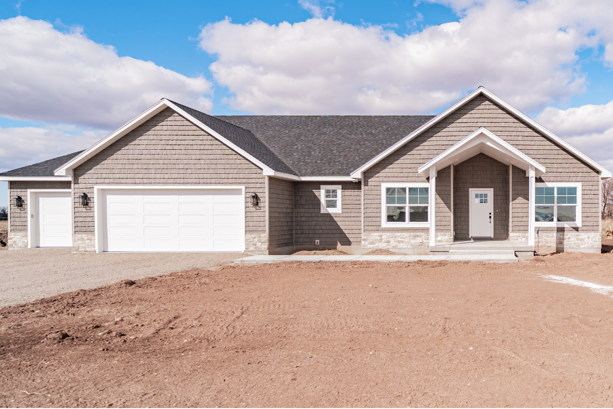 Exterior view of a newly constructed single-story home with gray siding, white trim, a three-car garage, and a covered front porch under a bright, cloudy sky.