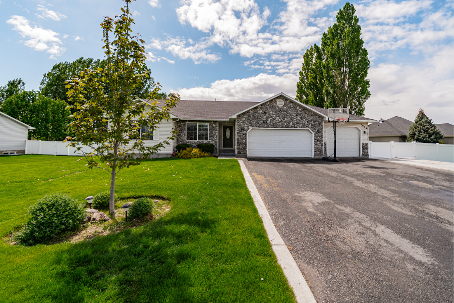 Single-story stone-front house with three-car garage in East Idaho — Spacious home with a mix of stone and siding exterior, large driveway, and landscaped front yard under a partly cloudy sky.