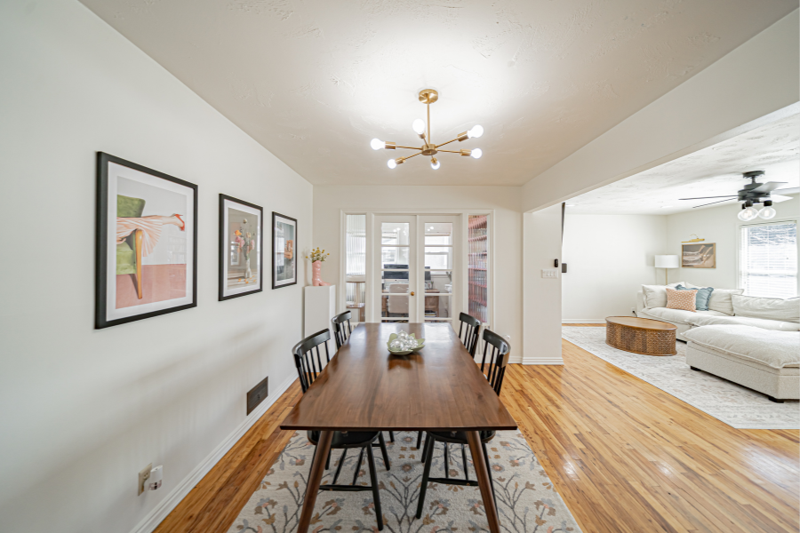 Modern dining room with mid-century lighting, hardwood floors, wall art, and an open view into a bright home office and adjacent living area.