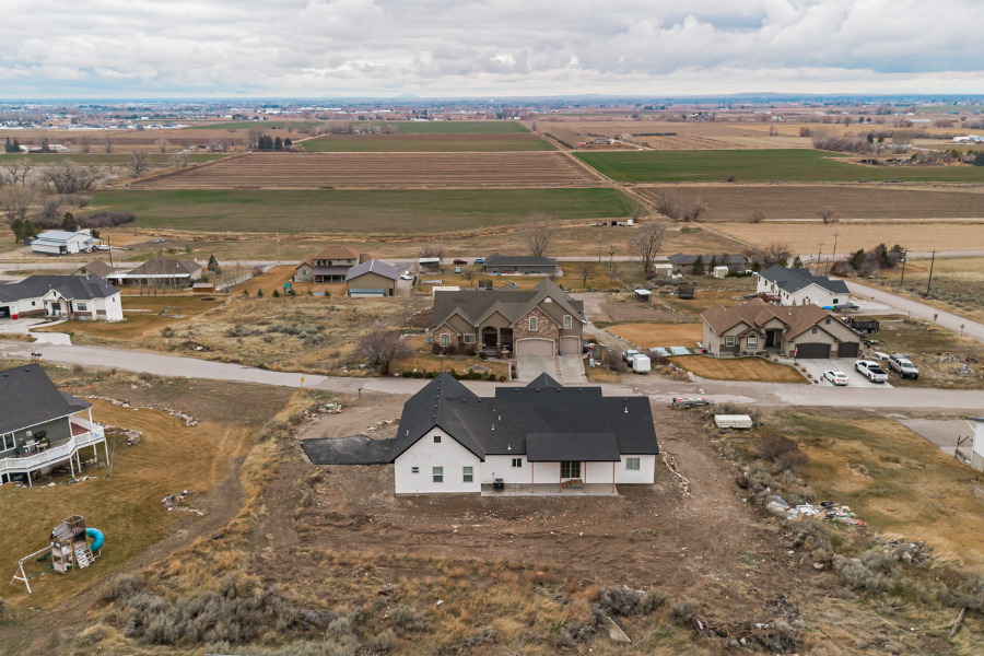 Drone view of a rural Idaho Falls property with custom homes, large lots, and expansive agricultural fields under a cloudy sky.