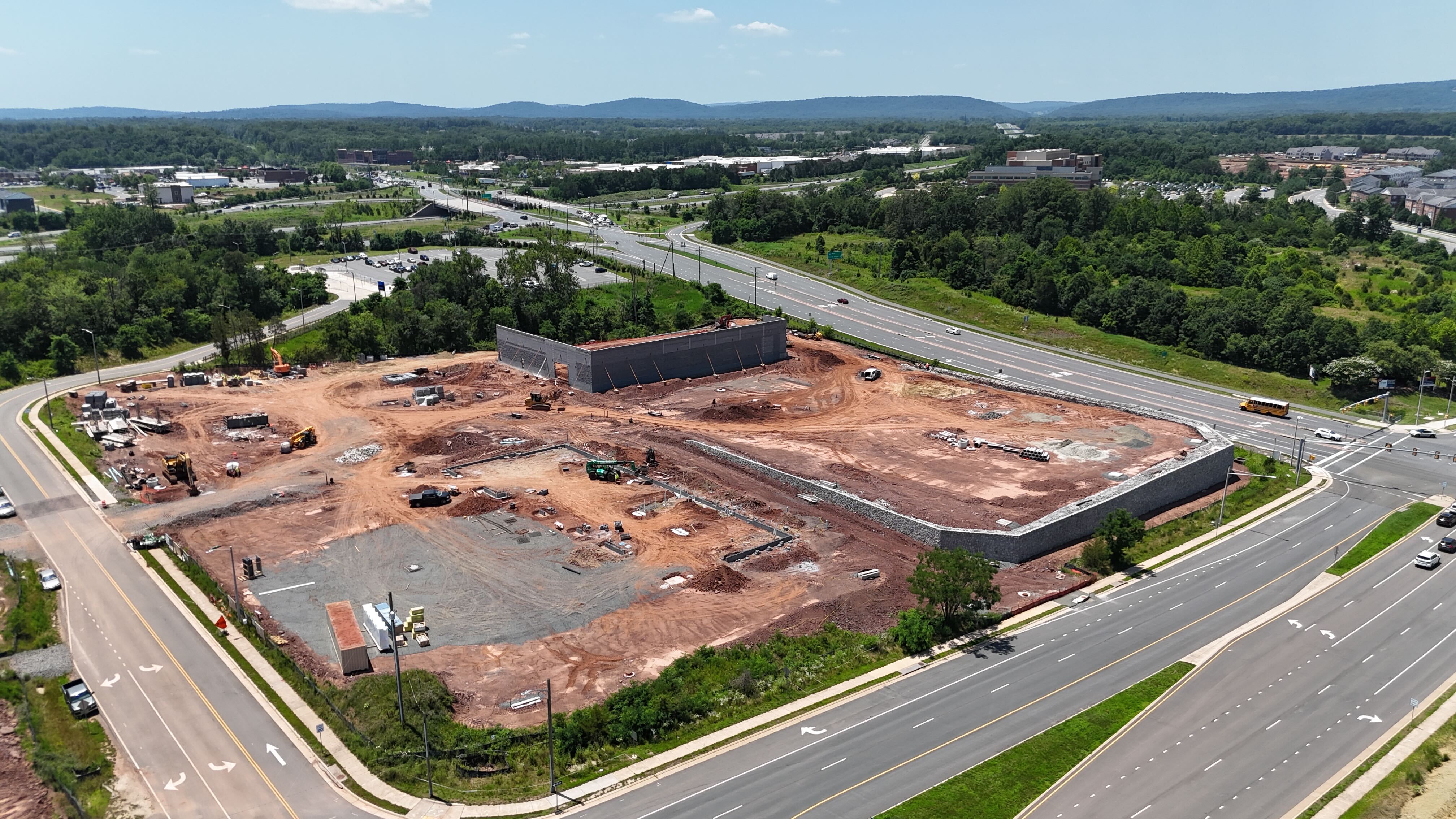 A wide aerial view of a large construction site in Haymarket, Virginia, with foundation walls in place, located next to a major highway, with the Bull Run Mountains in the background.