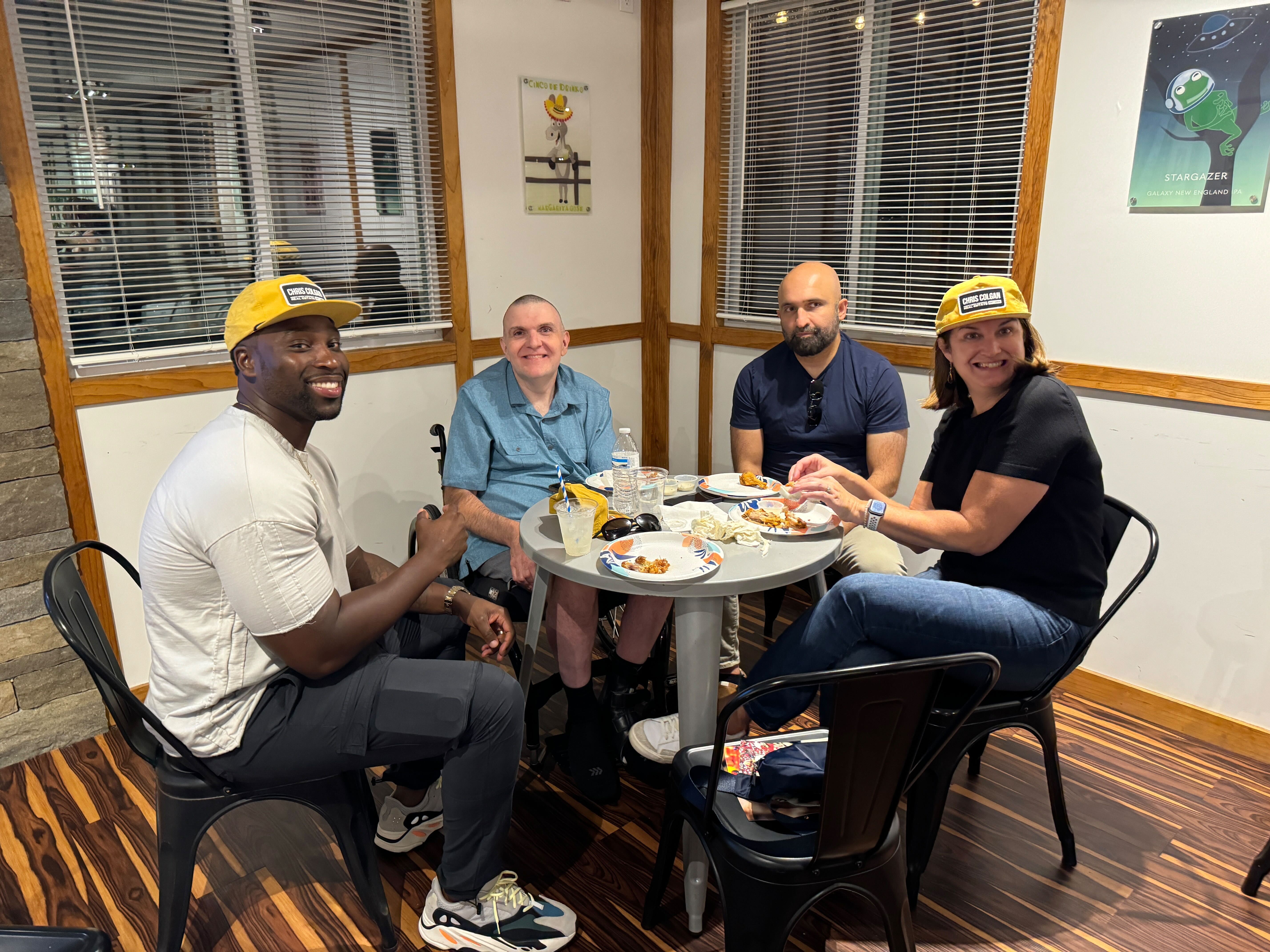 Four friends, some with yellow hats, smiling and enjoying a social gathering at a table inside The Farm Brewery at Broad Run in Gainesville, Northern Virginia.
