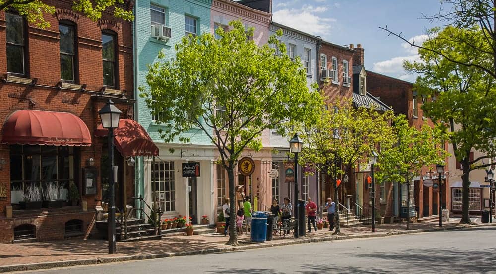 A sunny day on a commercial street in Old Town Alexandria, with visitors strolling past historic storefronts, boutiques, and restaurants.