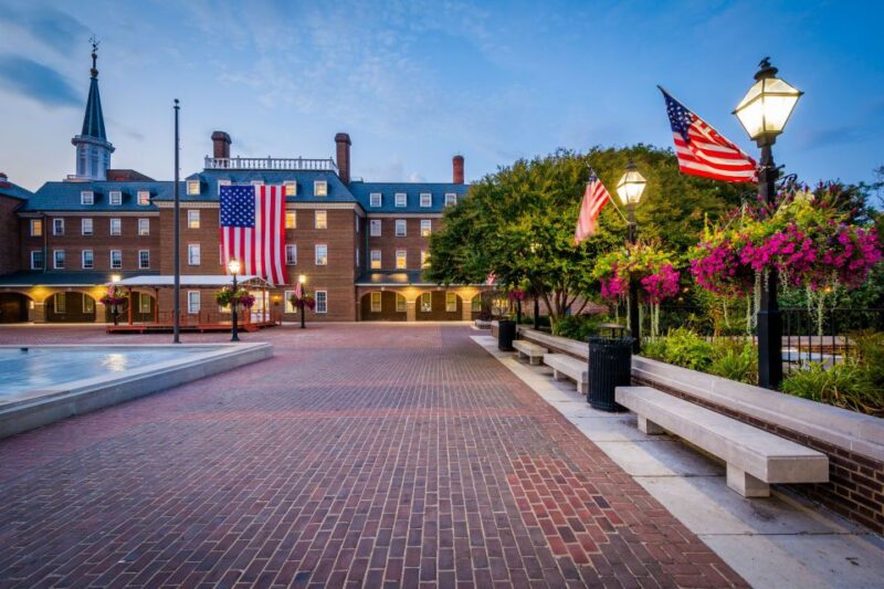 The historic brick plaza of Market Square in Old Town Alexandria at dusk, with glowing street lamps and an American flag on a colonial-style building.
