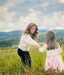A warm spring scene of a mother and daughter at a vineyard picnic in Virginia, holding flowers and wine glasses, with rolling hills in the background.