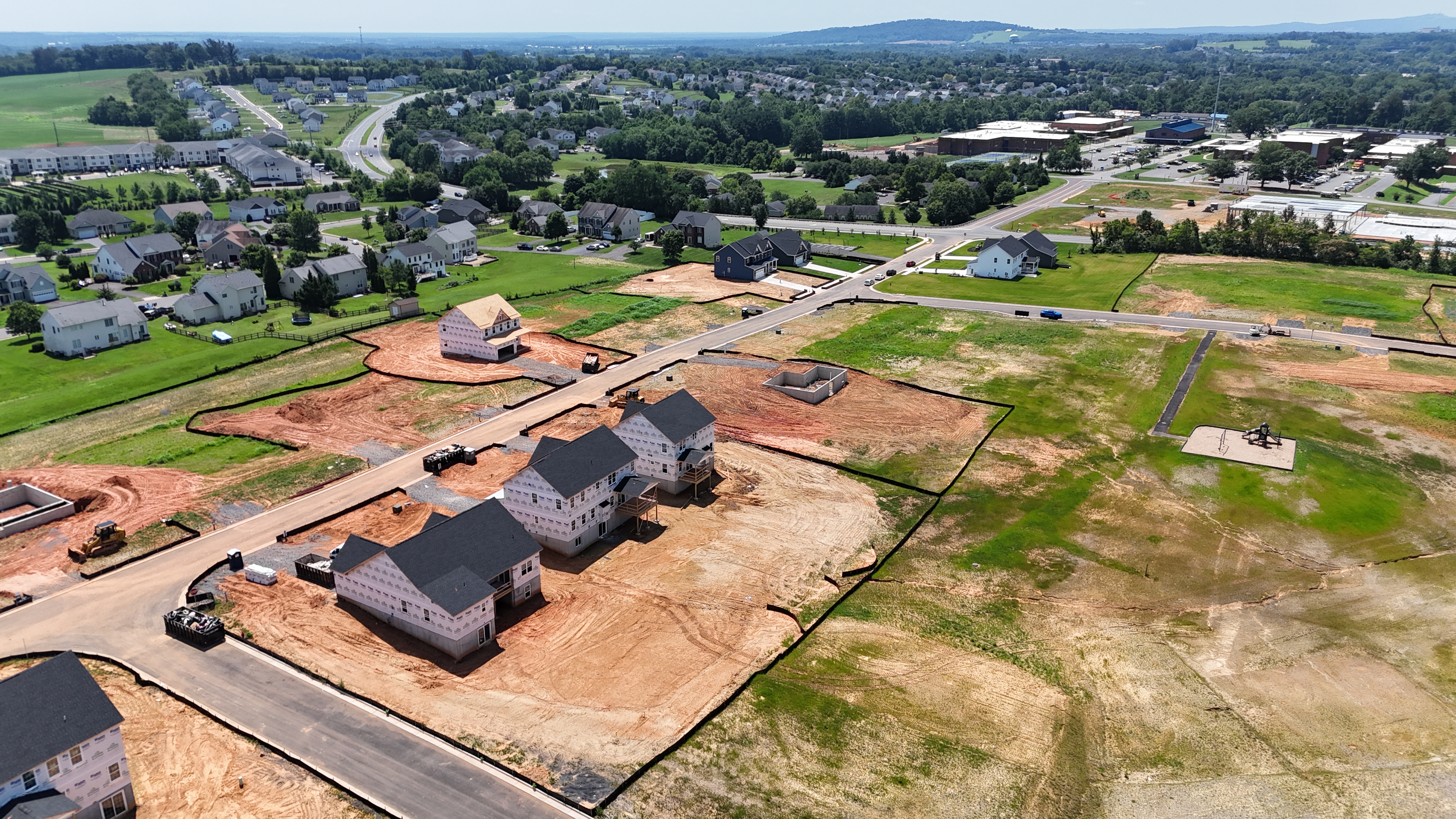 An aerial view of a new housing development under construction, showing several homes in the framing stage on graded dirt lots, with a school and established neighborhoods in the background.