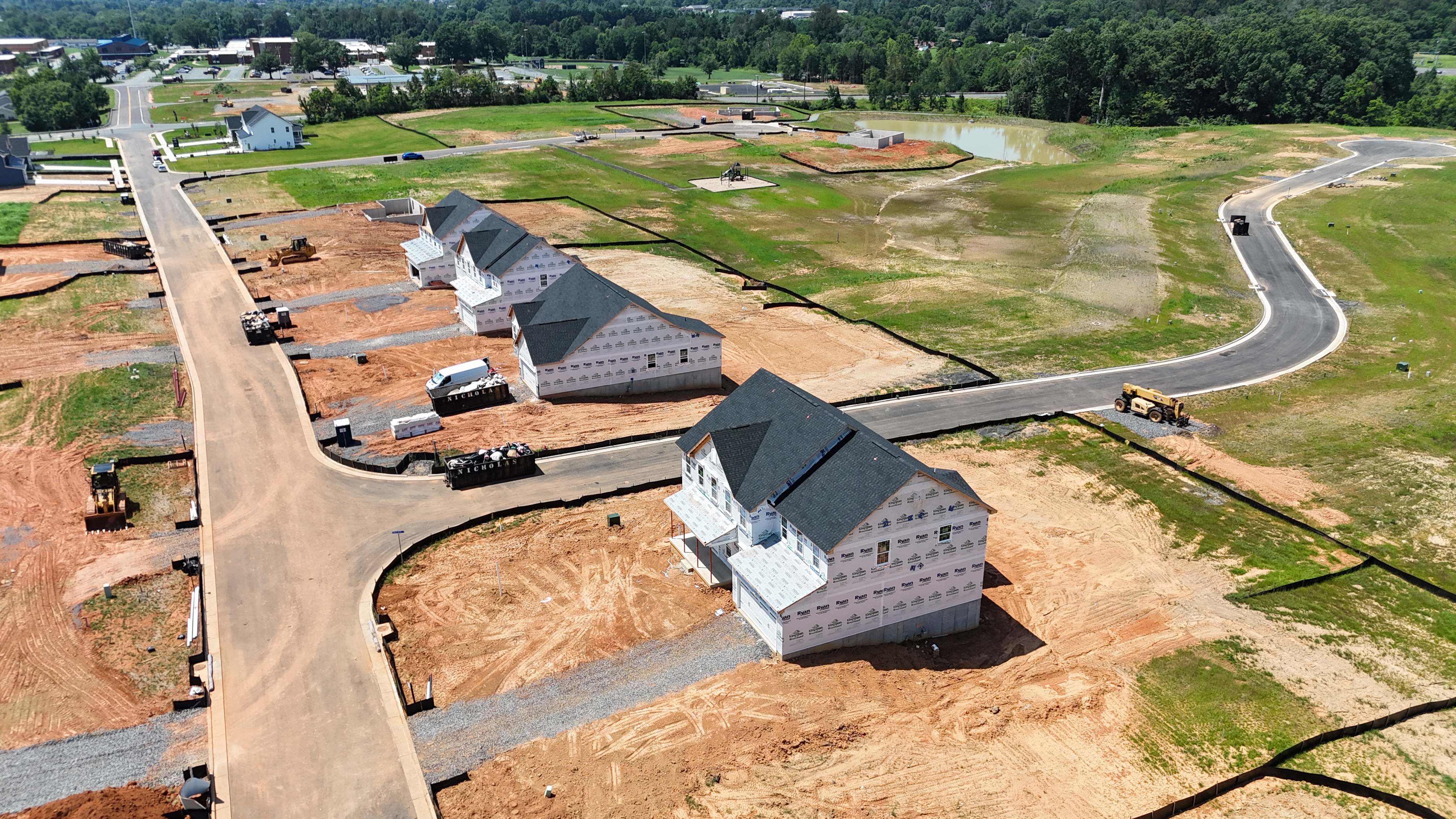 An aerial view of a new suburban housing development, showing a row of homes in the framing stage along a newly paved road, with construction equipment on the dirt lots.