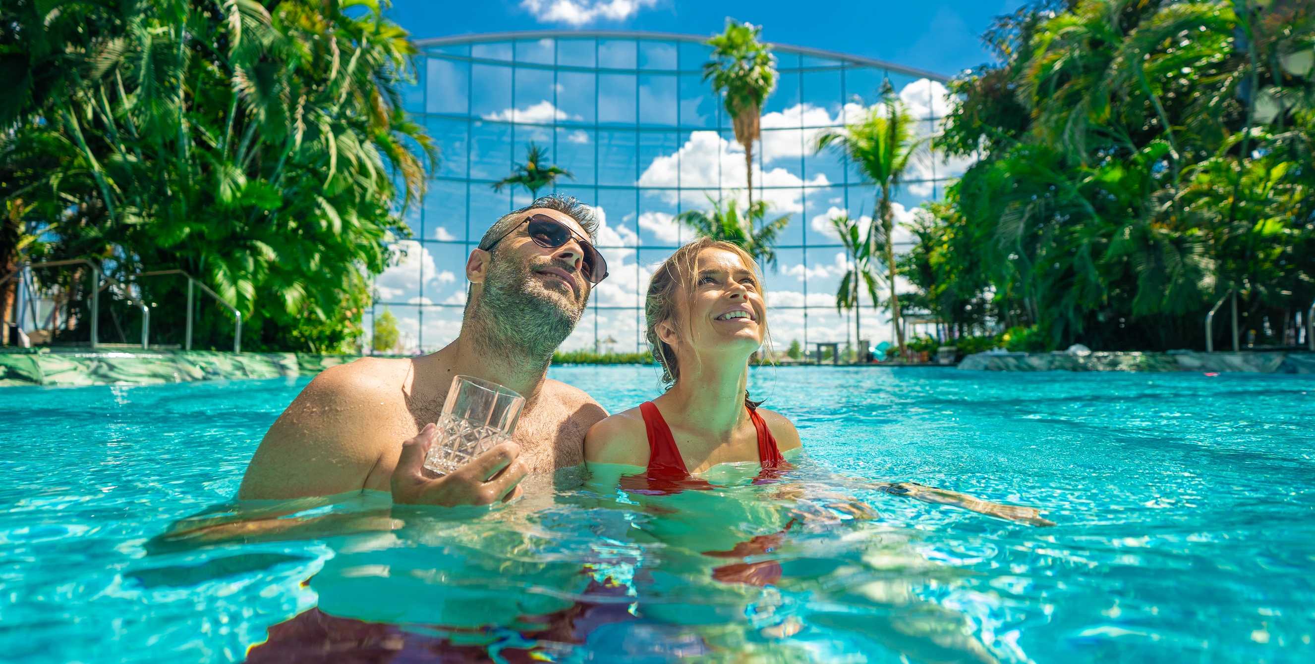 Smiling couple relaxing in a sunny blue pool at a Therme wellness resort, surrounded by tropical plants and a glass ceiling.