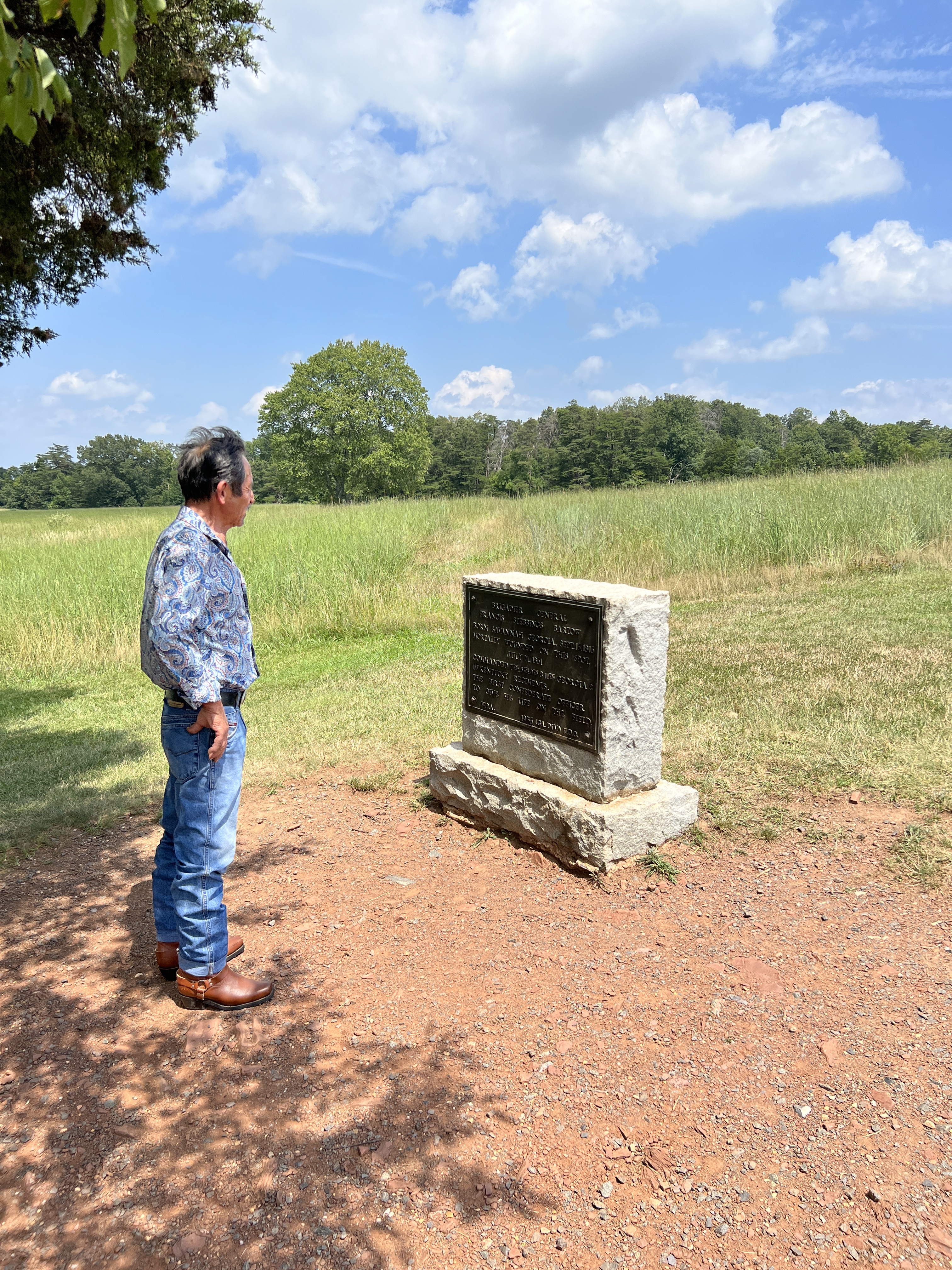 A visitor in a paisley shirt and jeans stands on a dirt path reading a bronze historical plaque on a stone monument at Manassas National Battlefield Park.