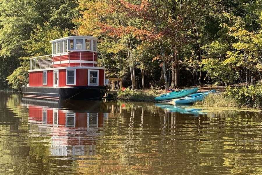 The red luxury tugboat houseboat Airbnb docked on a private lake in Louisa, Virginia, with kayaks on the shore and autumn leaves on the trees.