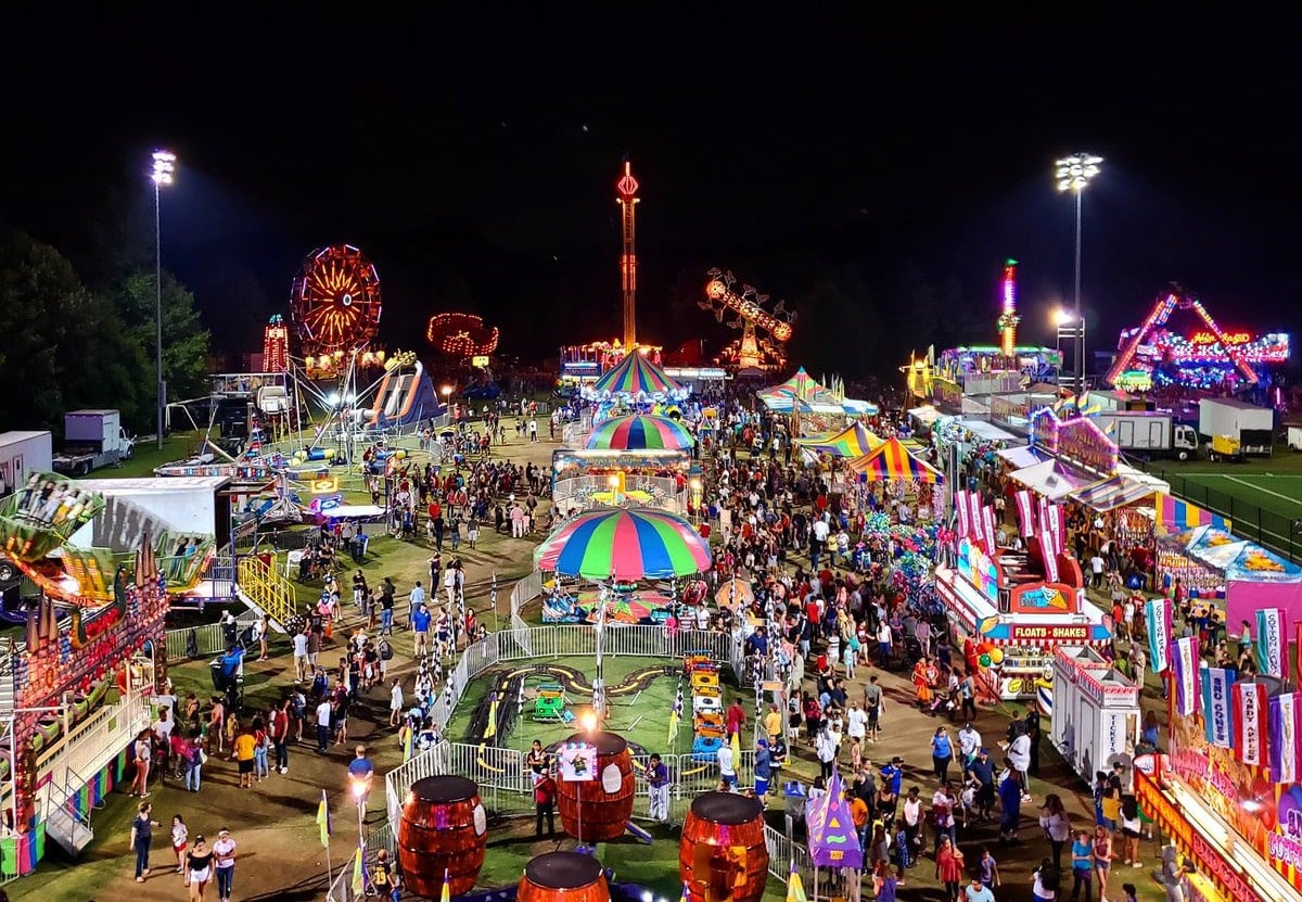 A wide, elevated view of the Arlington County Fair at night, bustling with crowds of people among the brightly lit carnival rides, with a Ferris wheel glowing against the dark sky.