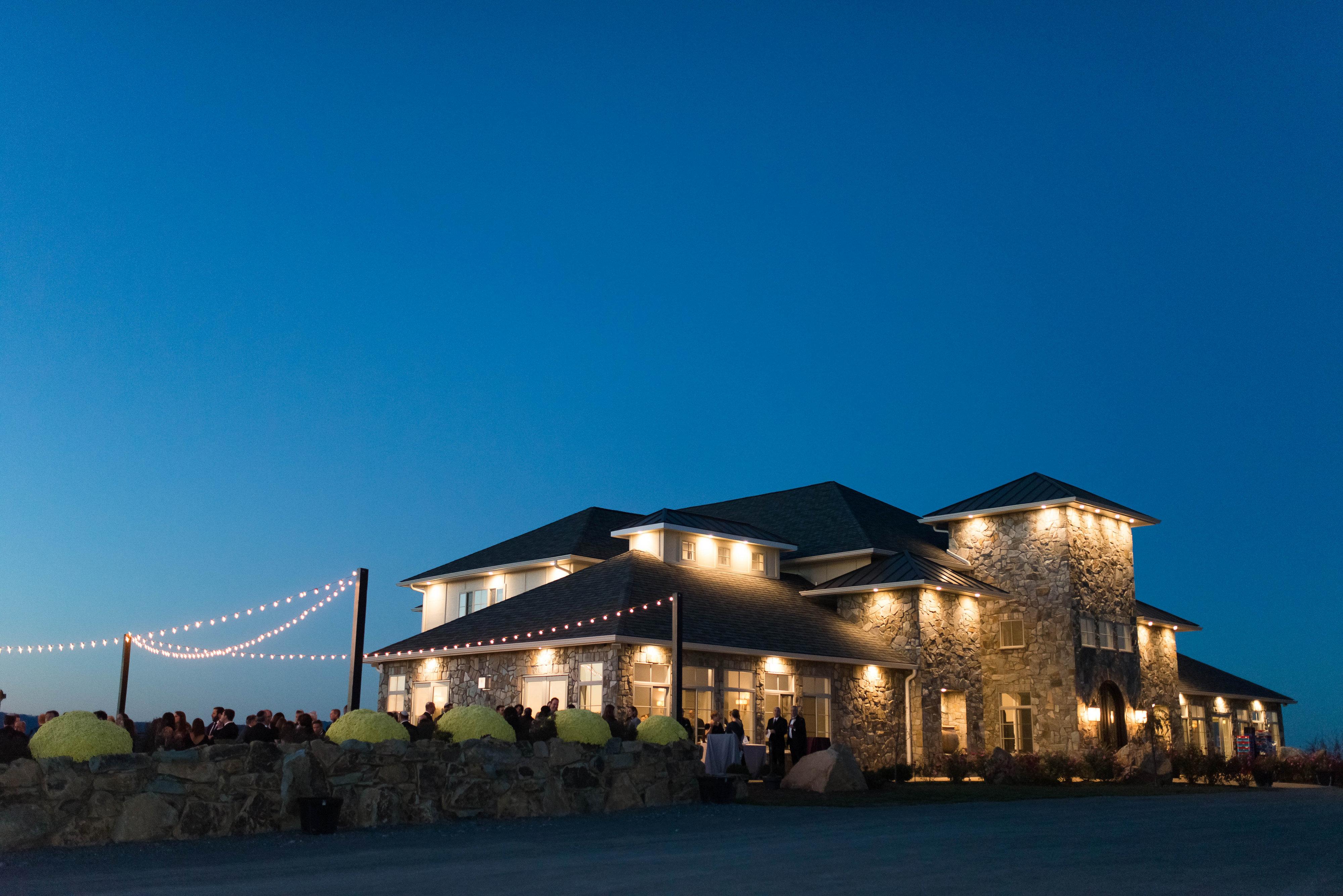 An evening wedding reception at Blue Valley Vineyard and Winery, showing the stone-facade venue illuminated against a deep blue dusk sky, with guests gathered on the patio under strings of glowing cafe lights.