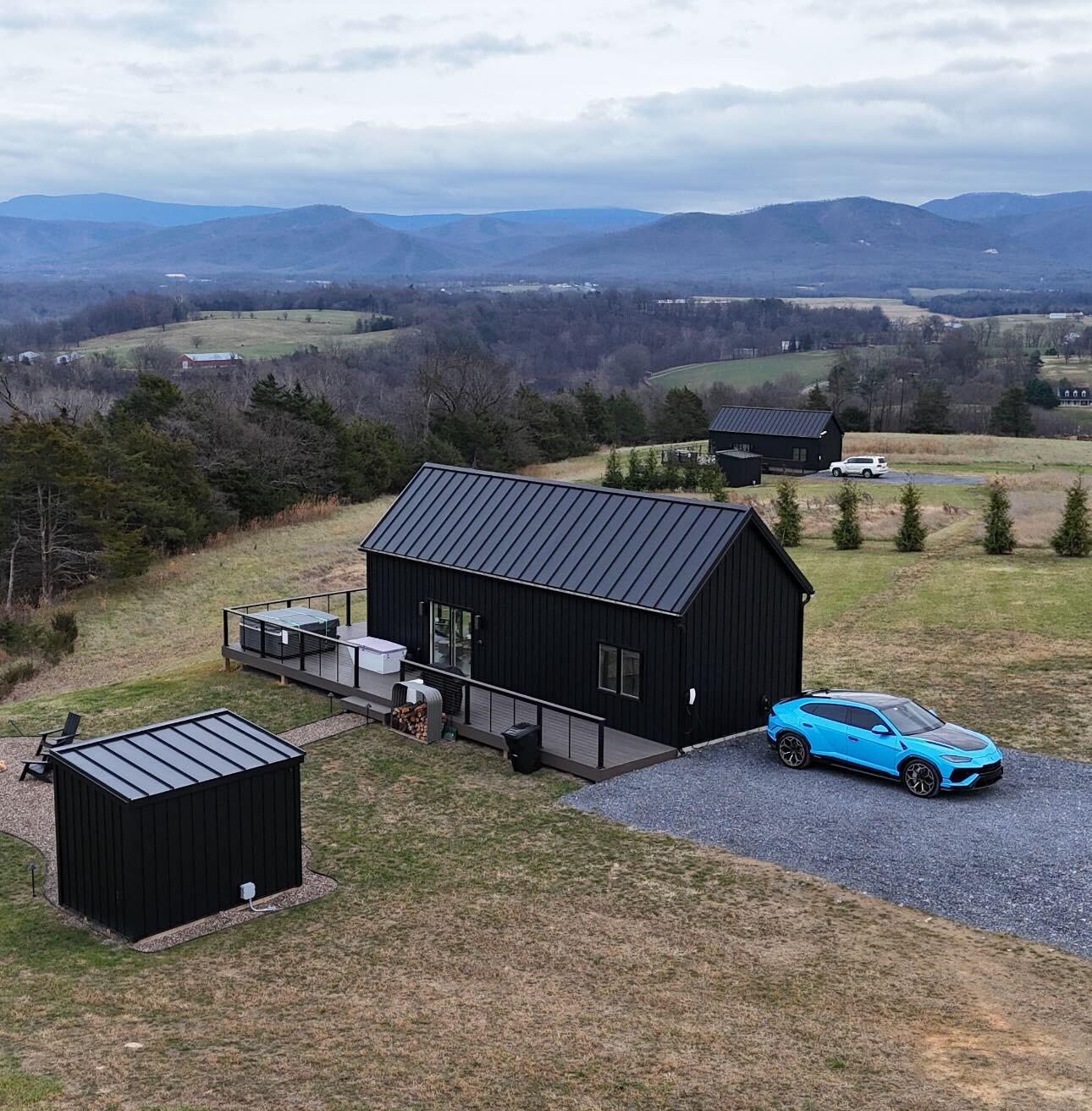 A bright blue Lamborghini Urus parked on a gravel driveway in front of a modern black cabin with a deck and hot tub, overlooking the Blue Ridge Mountains in Virginia.