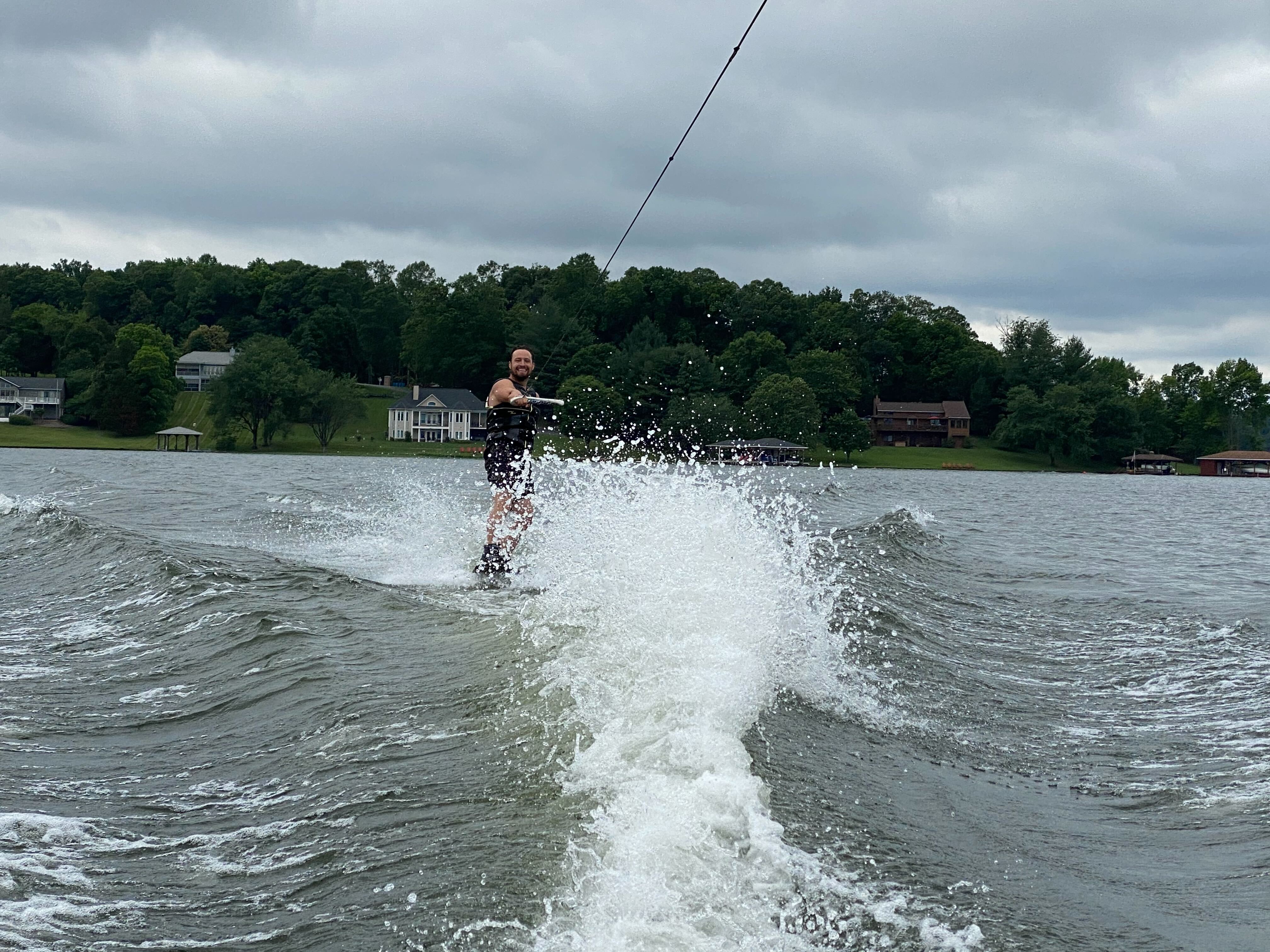 A man wakeboarding on Lake Anna in Virginia, with a large spray of water behind him and the tree-lined shore in the background.