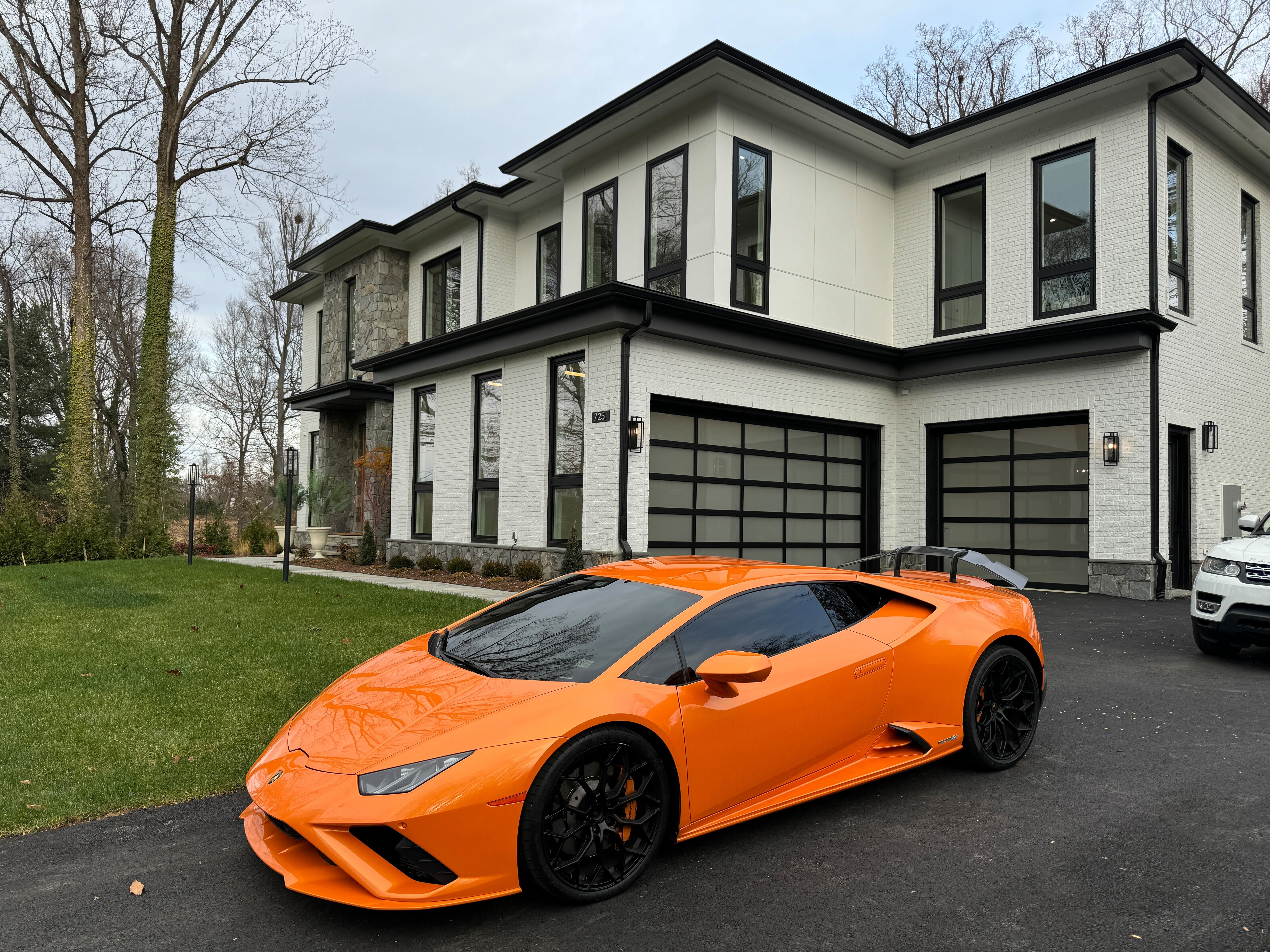 An orange Lamborghini Huracan parked in the circular driveway of a large, modern luxury home with a white brick and stone exterior in Great Falls, Virginia.