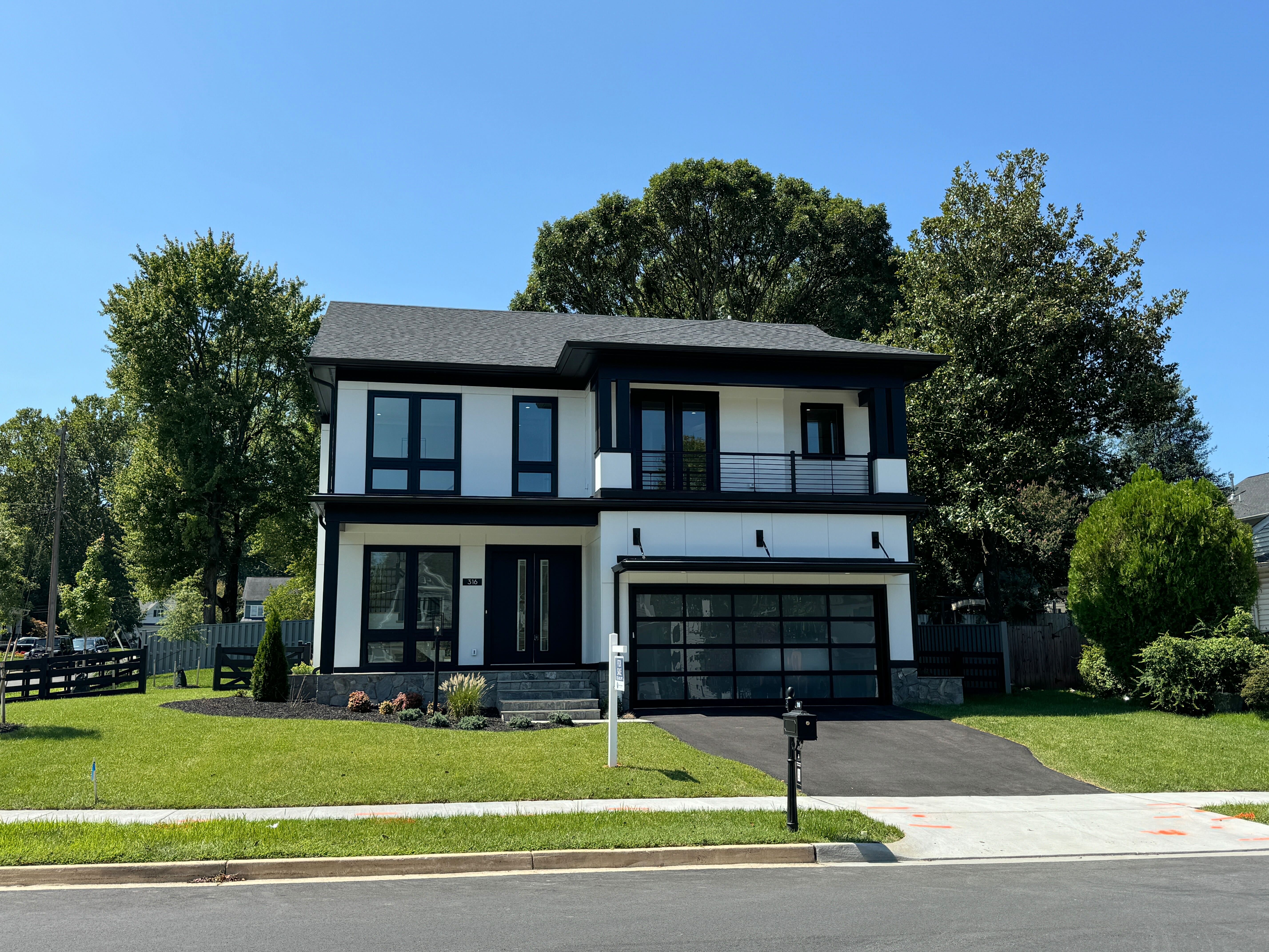 The exterior of a modern two-story new construction home with a white facade, black trim, a second-floor balcony, and a glass-paneled garage door.