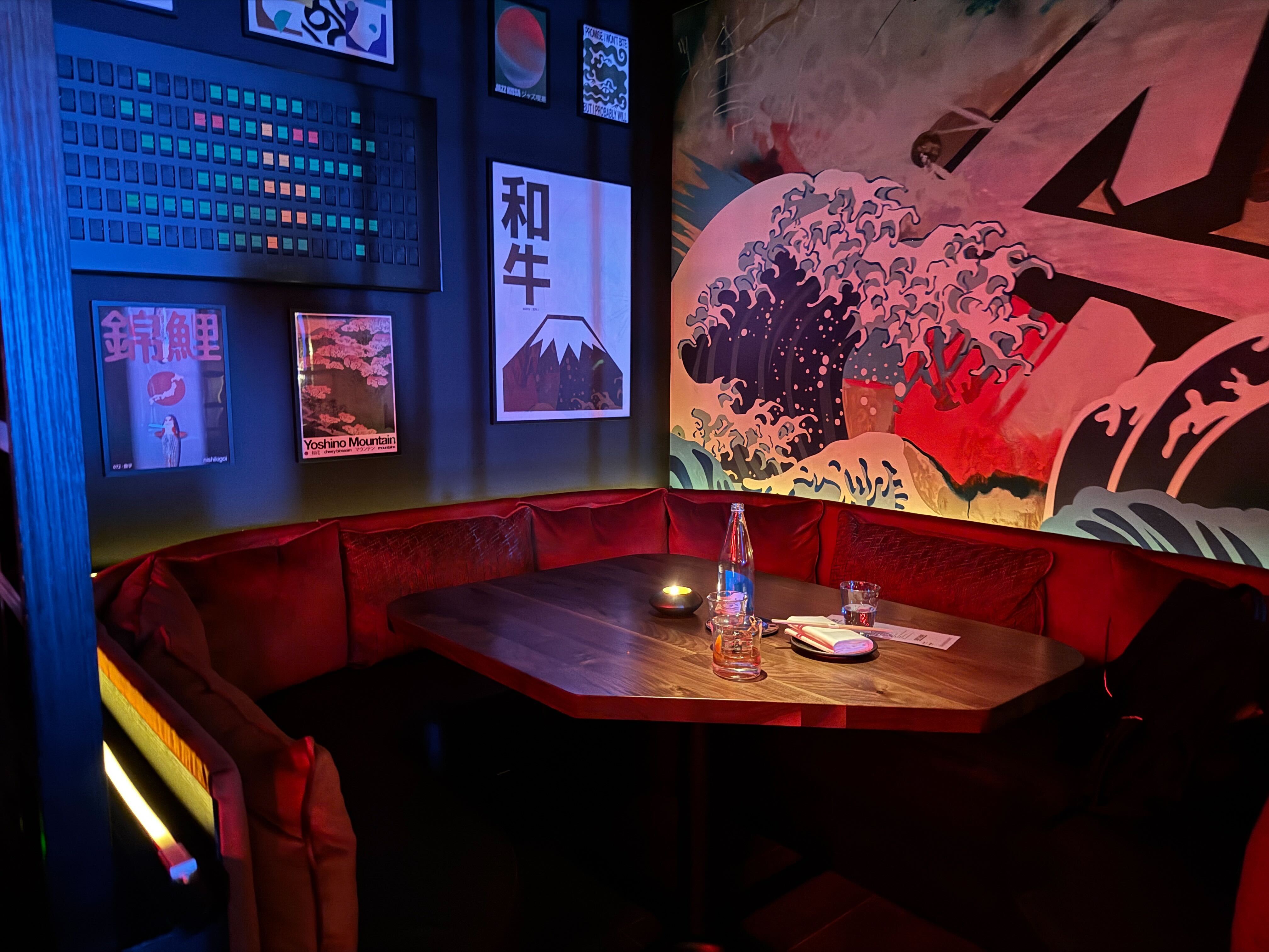 A stylish, dimly lit corner booth in a Japanese restaurant in Washington, D.C., featuring red velvet seating and a large mural inspired by 'The Great Wave off Kanagawa'.