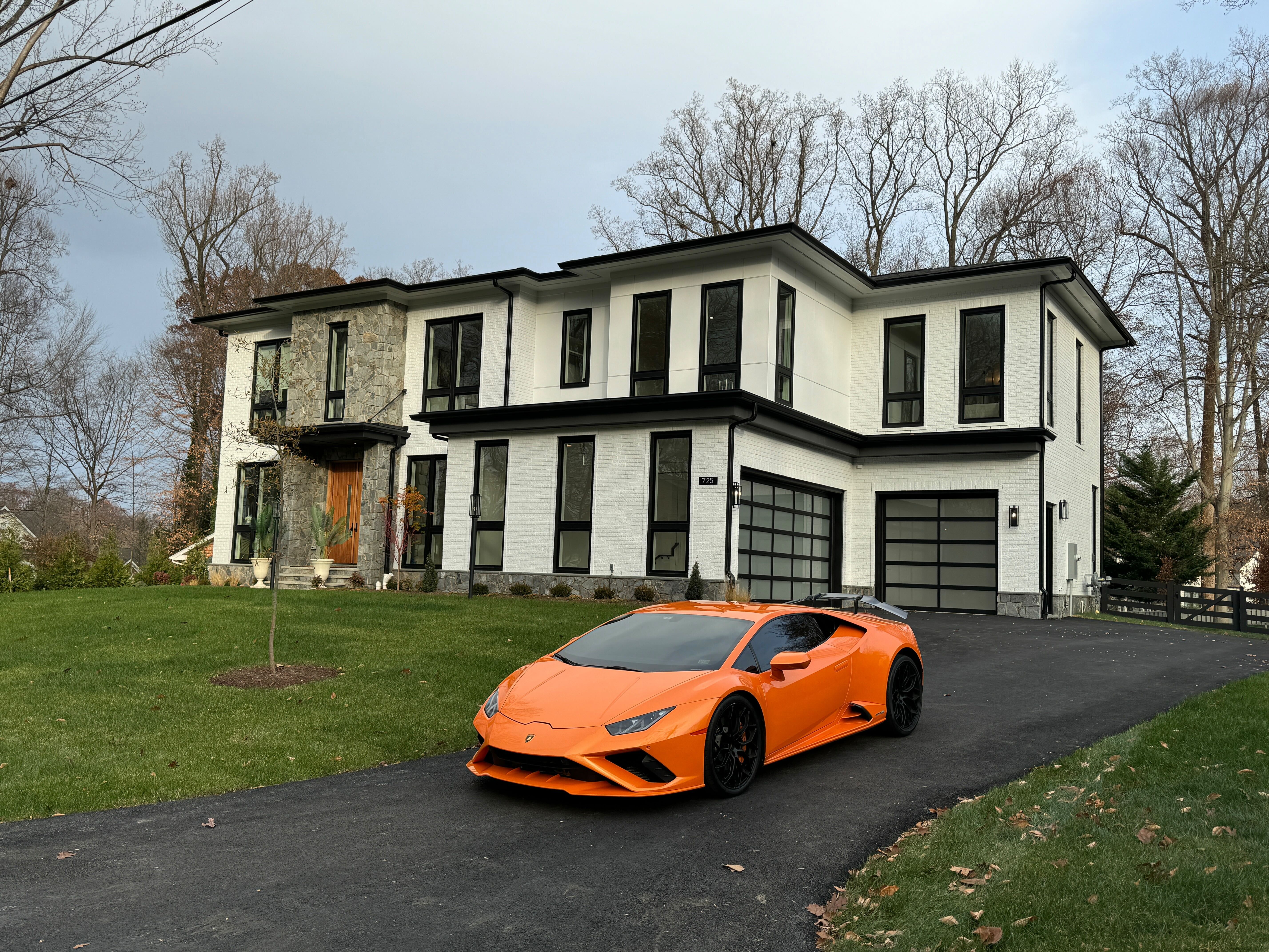 A bright orange Lamborghini Huracan parked in the driveway of a large, modern house with white brick and stone accents.