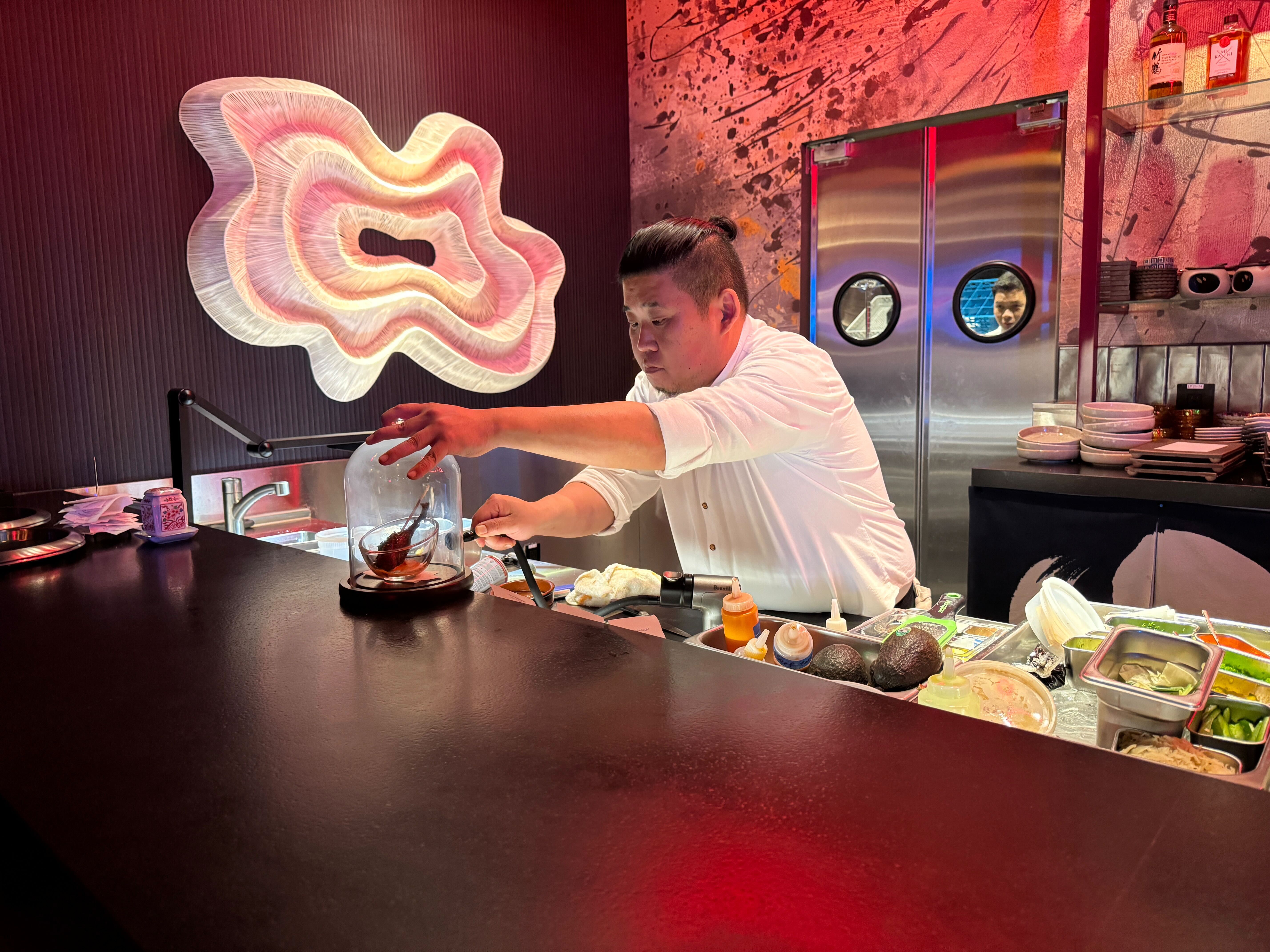 A sushi chef at an omakase restaurant carefully places a glass cloche over a dish, with a modern art piece on the wall behind him.