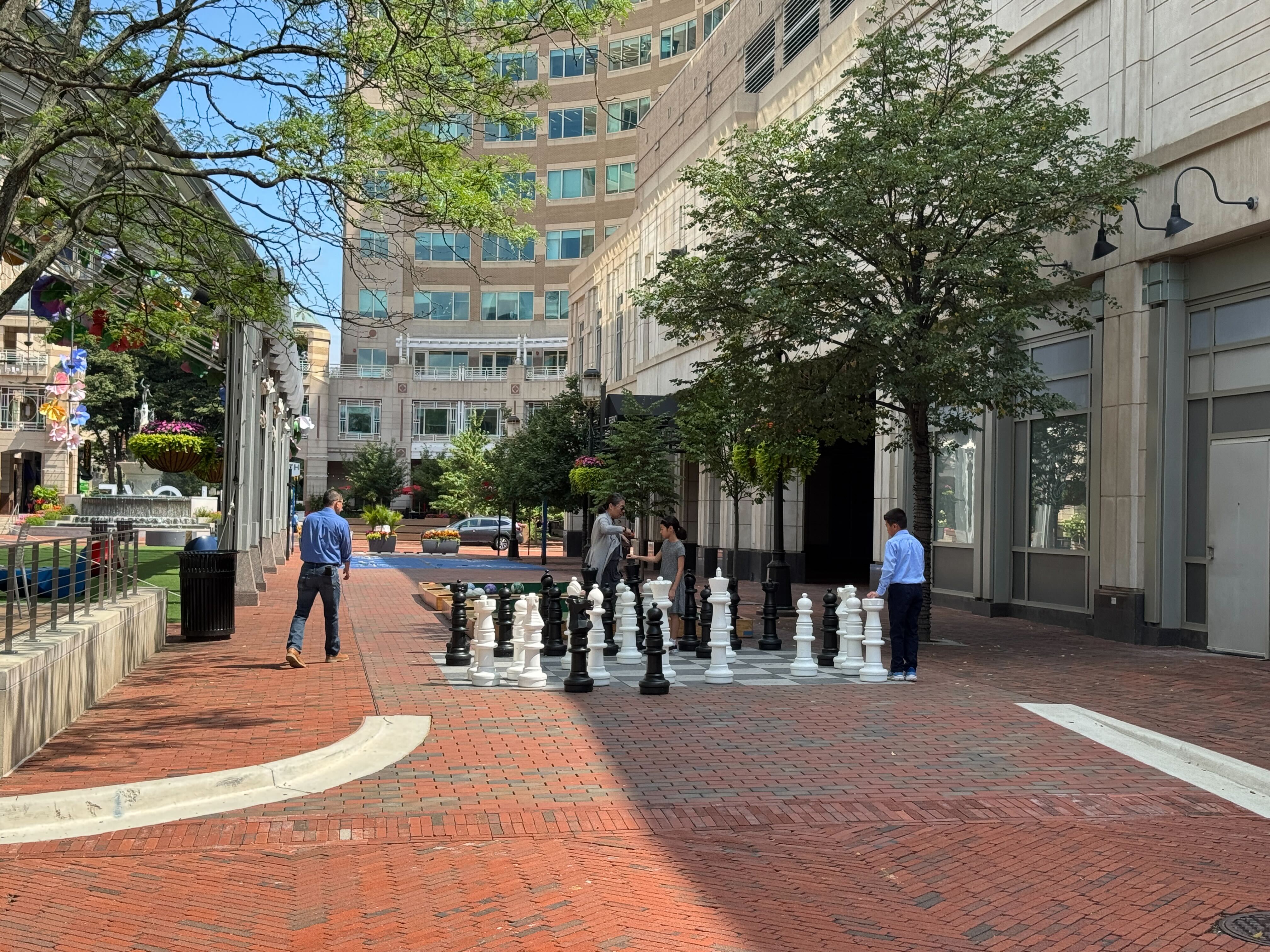 People playing on a giant outdoor chessboard on the brick plaza of Reston Town Center, surrounded by trees and modern office buildings.