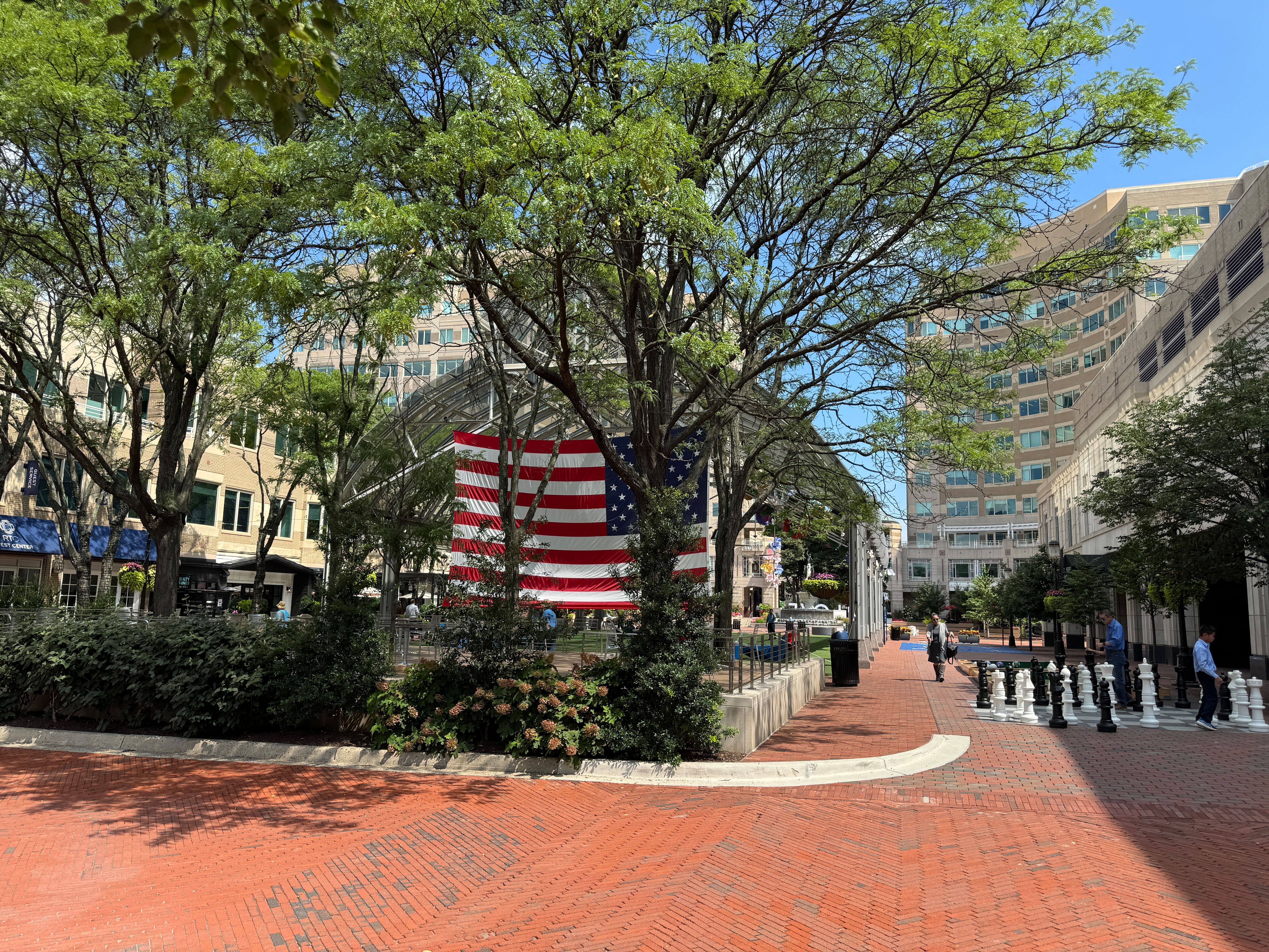 A large American flag hangs between trees over the public plaza and giant chess set at Reston Town Center, a hub of community life in Reston, VA.
