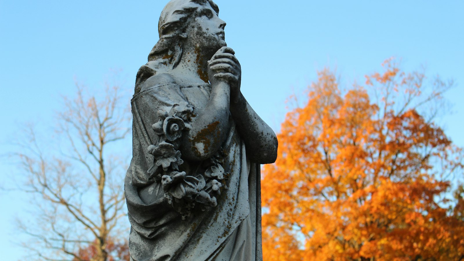 Weathered stone cemetery statue framed by bright orange autumn leaves under a clear blue sky