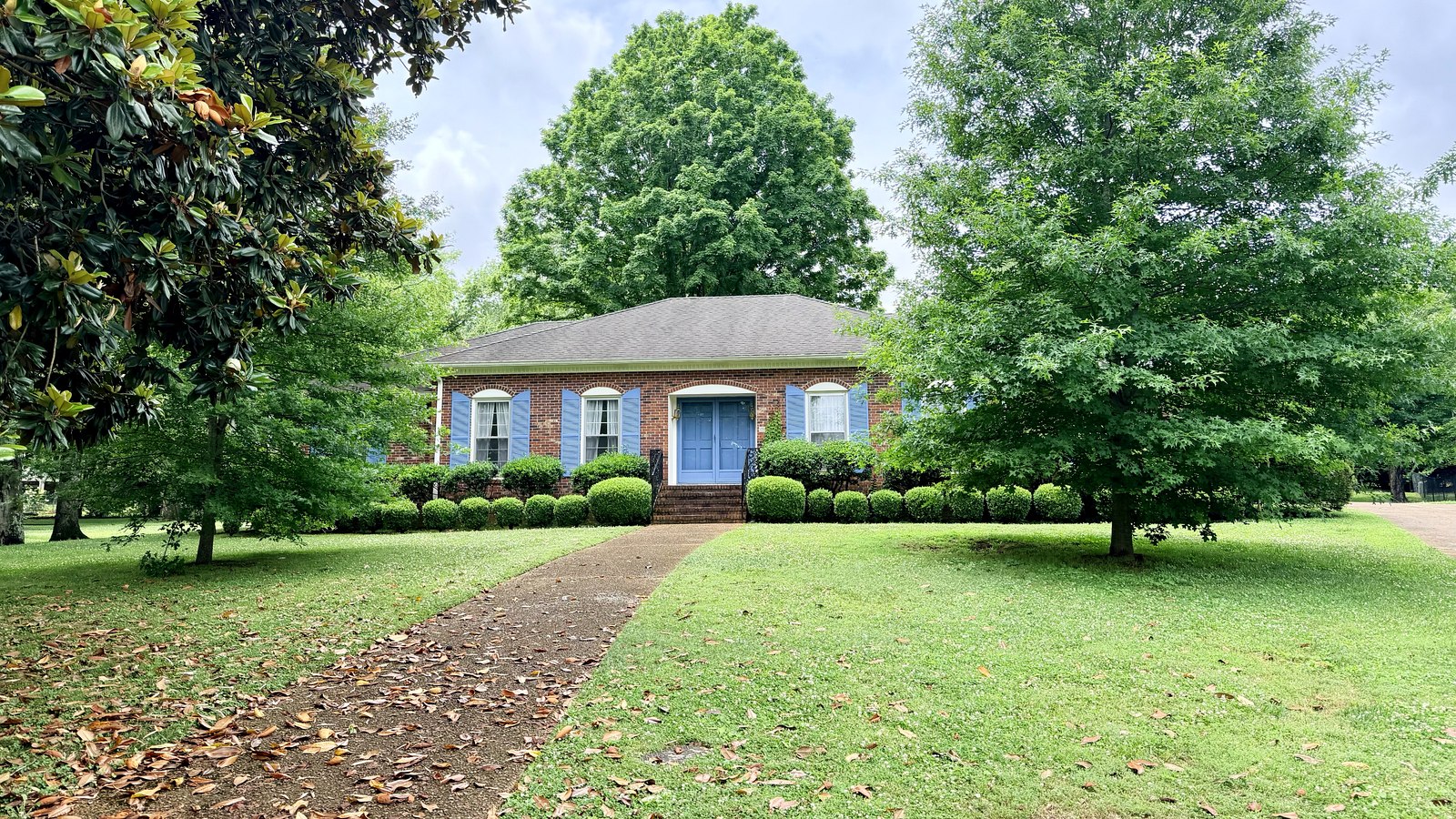 Brick ranch home with blue shutters, a clipped boxwood hedge and a walkway under mature shade trees in Jones Valley, Huntsville, Alabama
