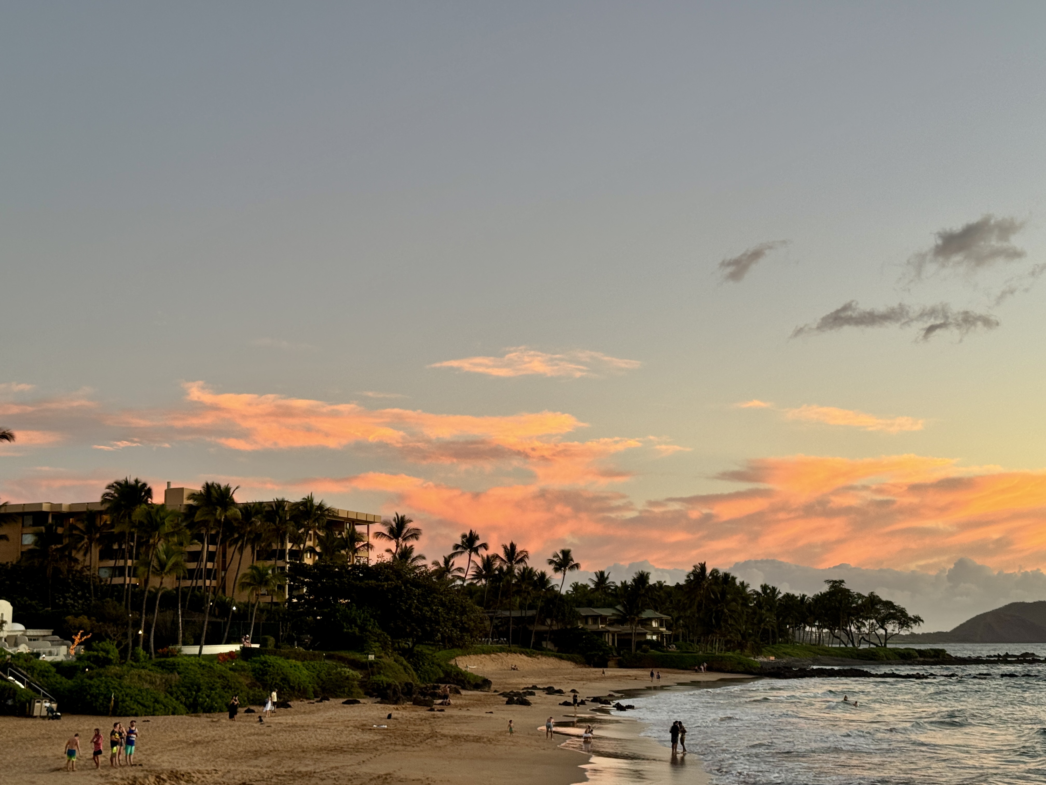 Scenic Wailea beach at sunset with soft waves, palm trees, and a view of Molokini Crater on the horizon