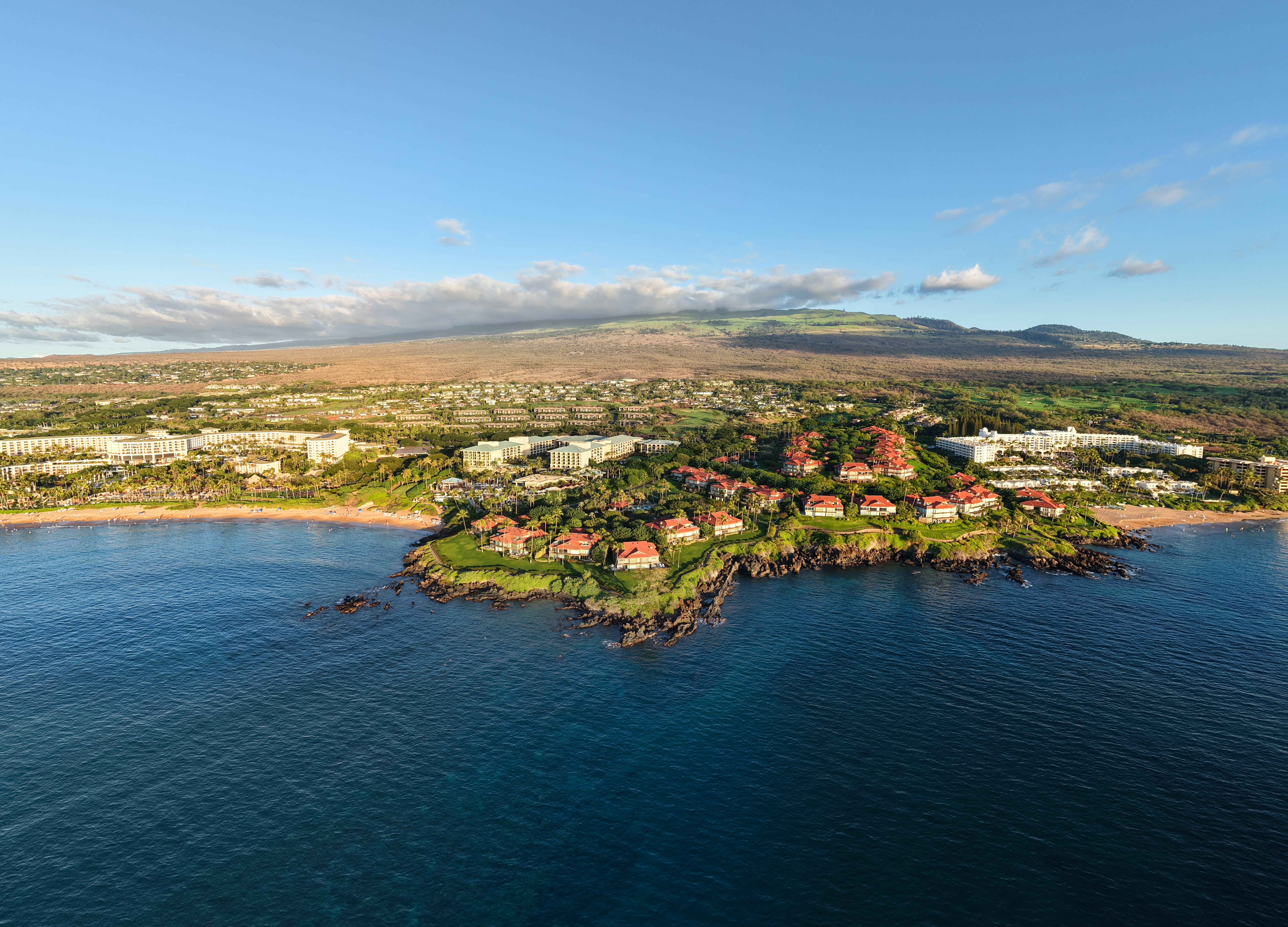 Aerial view of the Wailea coastline on Maui's South Shore, showing Wailea Point luxury condominiums on a lava rock peninsula in the foreground, with the Grand Wailea resort and Ho'olei villas visible inland, the Four Seasons and Fairmont Kea Lani along the shoreline to the right, and Haleakalā rising above the resort corridor under a clear blue sky.