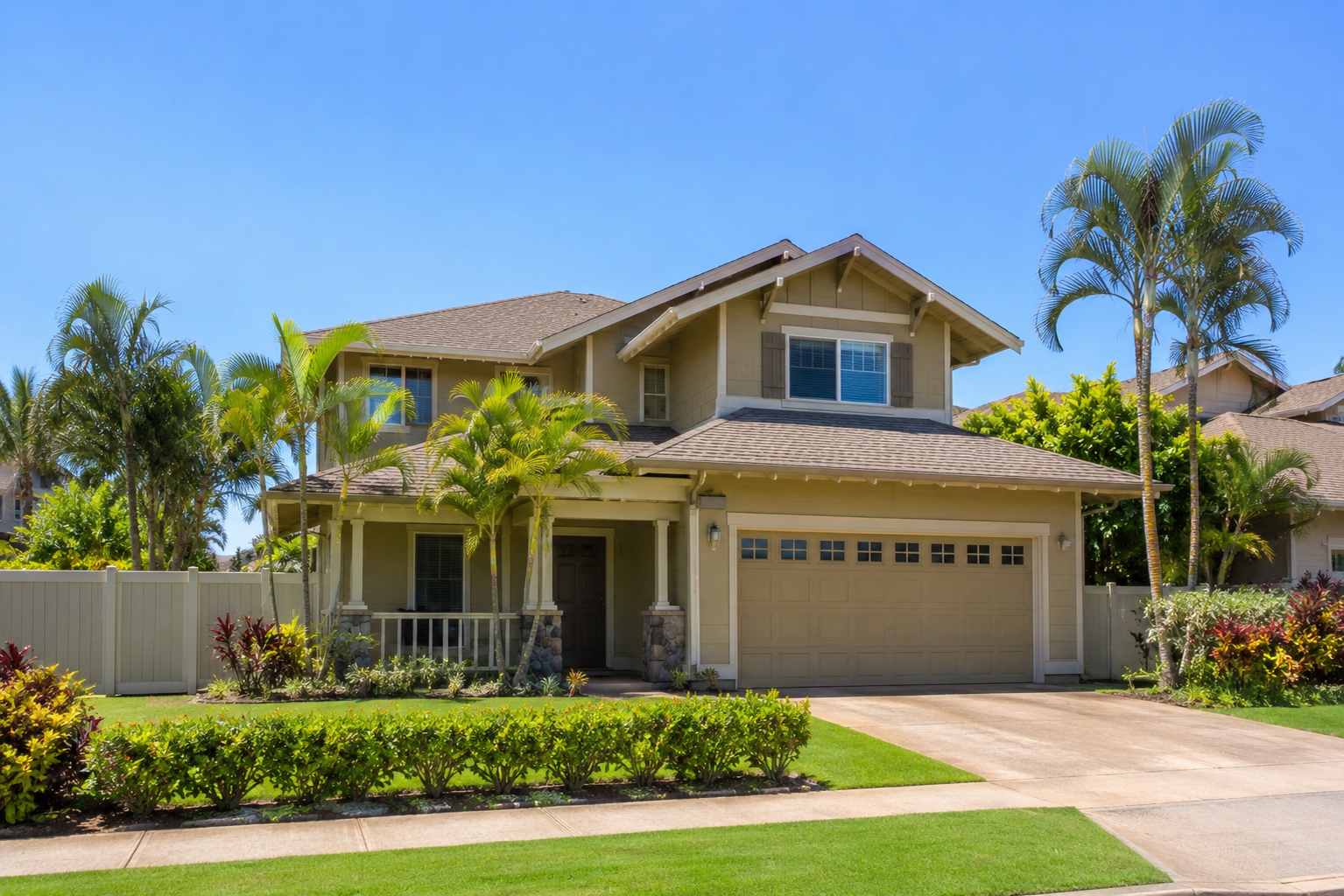 Two-story single-family home in Hale Piilani subdivision Kihei Maui featuring two-car garage, covered front porch, tropical landscaping, fenced yard, and classic Hawaii residential architecture under blue skies