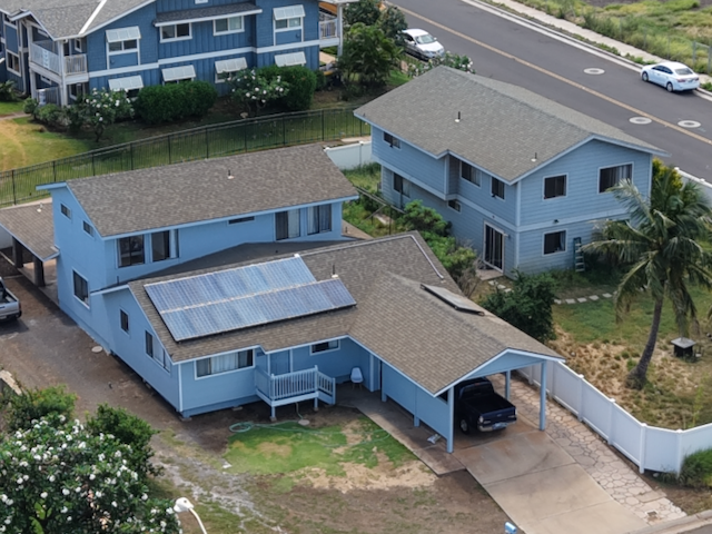 Front view of a two-story light blue home in North Kihei, with a covered porch, a solar-panel roof, and a fenced yard