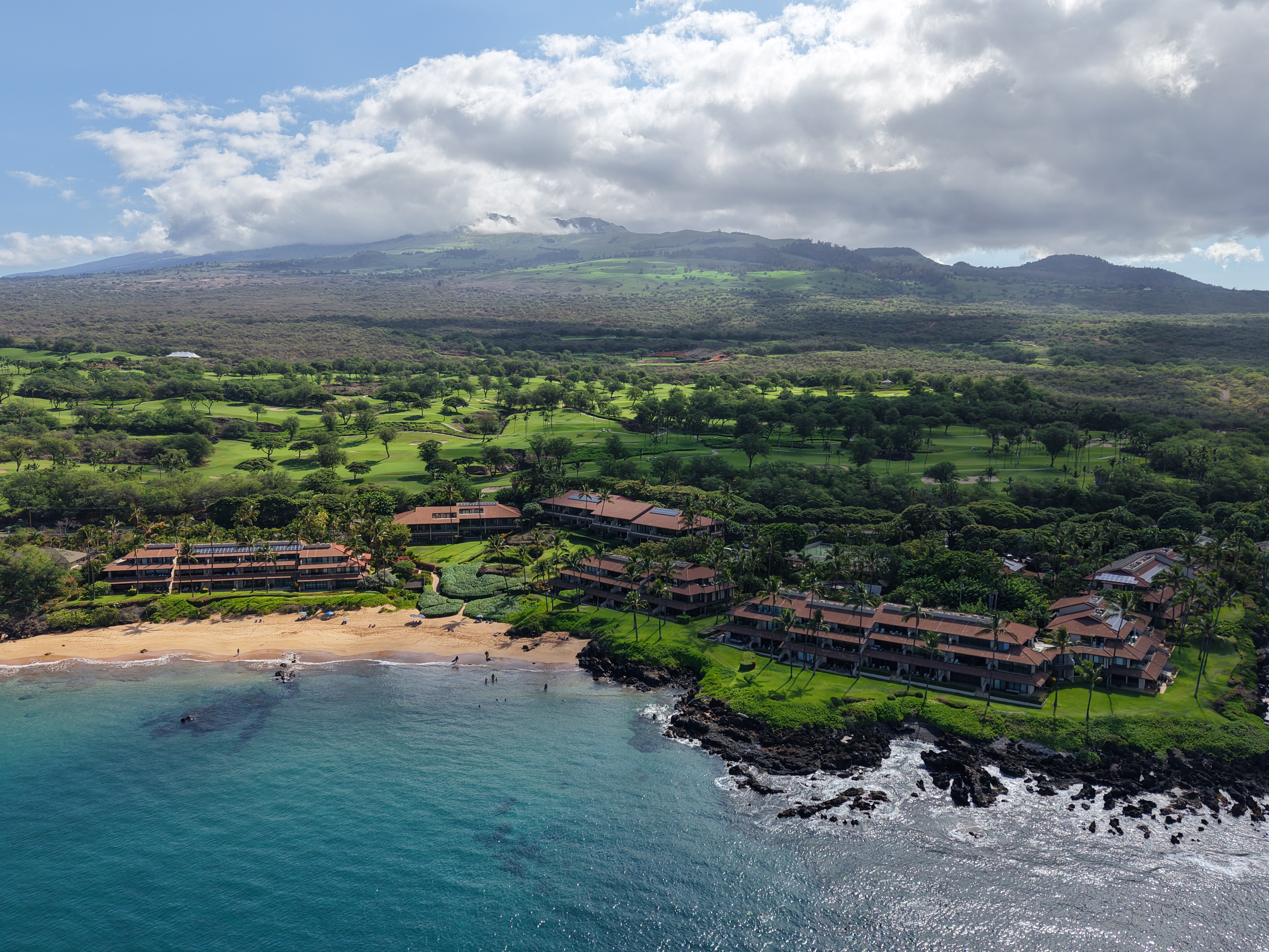 Aerial view of Makena Surf condominium complex on the South Maui coastline, showing low-rise beachfront buildings surrounded by lush tropical landscaping and palm trees, with a sandy beach, turquoise ocean water, volcanic lava rock shoreline, Makena Golf Course fairways, and the West Maui Mountains in the background.