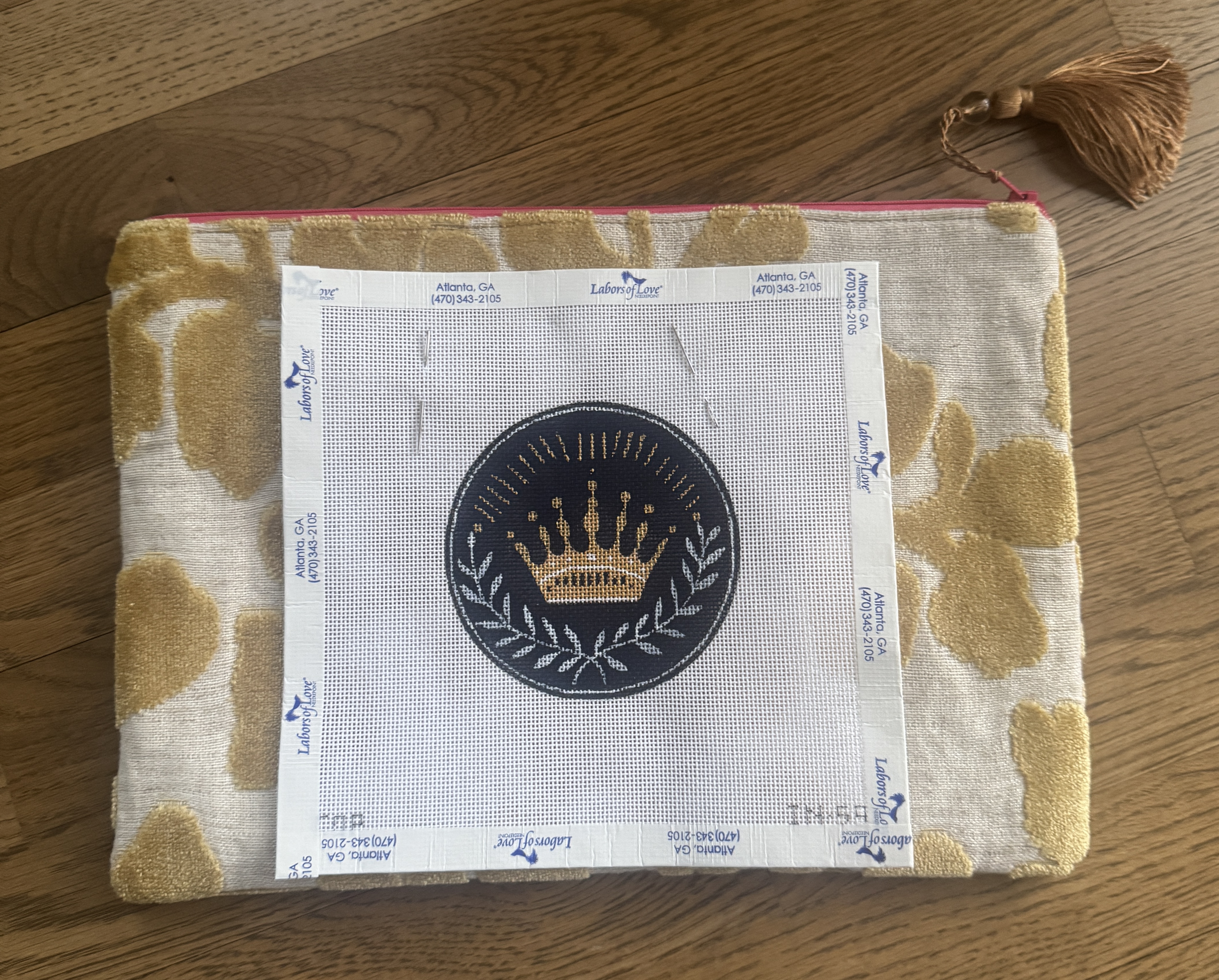 Needlepoint canvas featuring a gold crown inside a dark circular emblem, resting on a neutral zipper pouch with a gold tassel on a wood floor. The needlepoint piece was purchased during a trip to Atlanta.