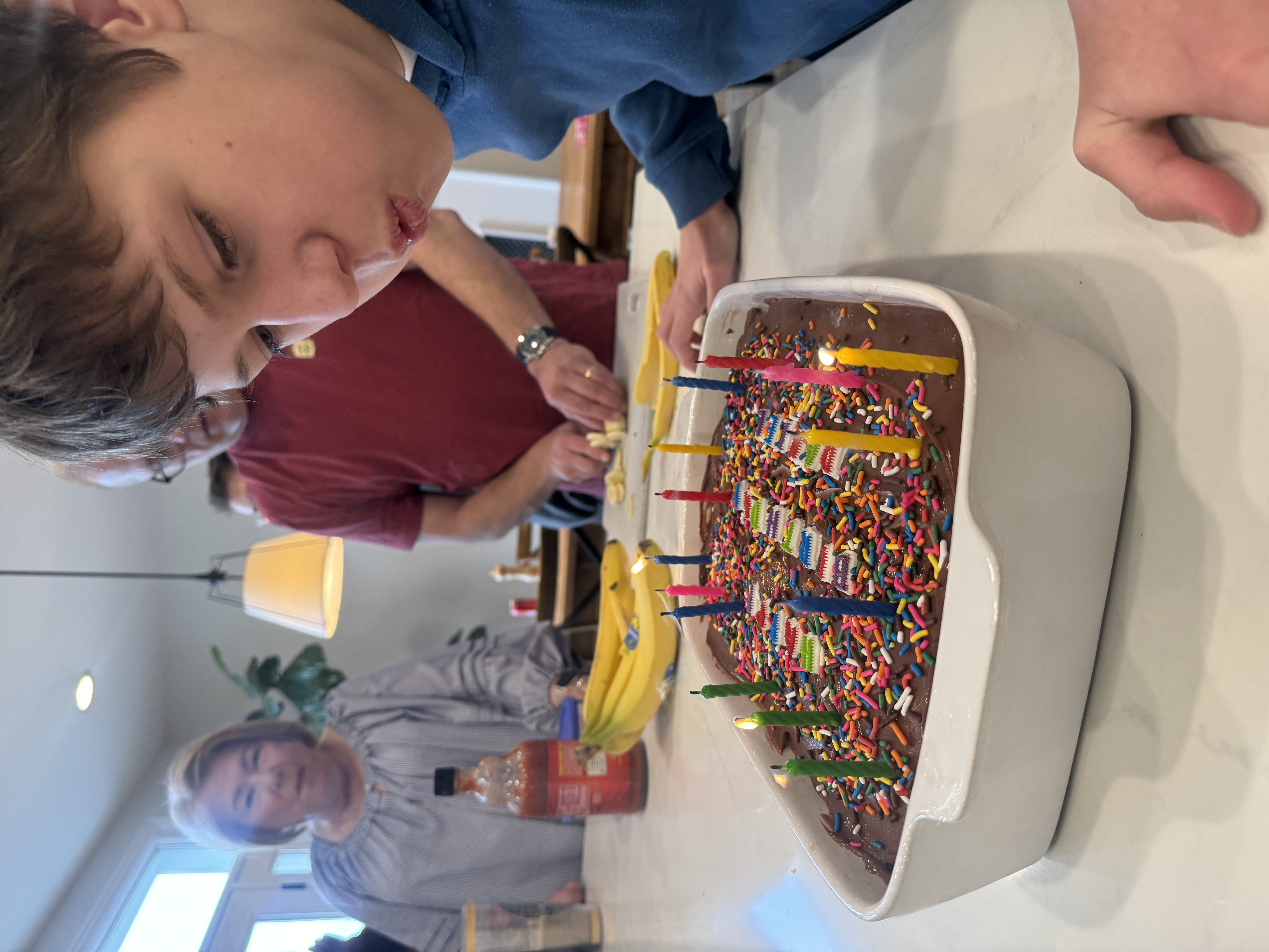 Daniel blowing out candles on his birthday ice cream cake while family gathers around the kitchen island.