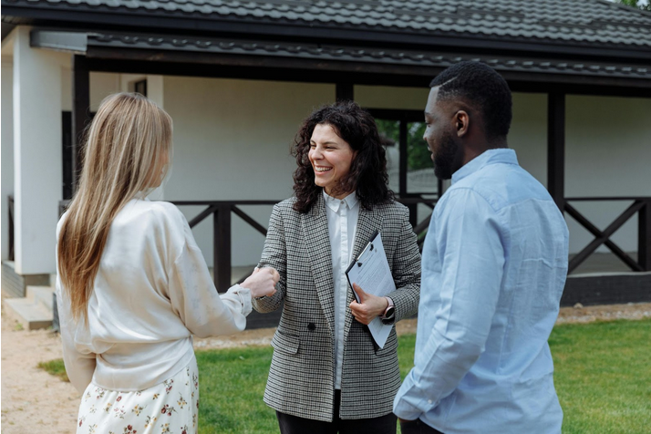 A real estate agent shaking hands with a couple in front of a house, discussing home buying options.
