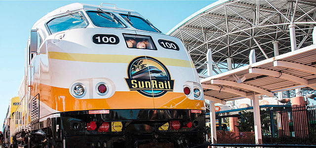 Close-up of a yellow and white SunRail locomotive numbered 100 at a station platform, with a modern canopy structure and blue sky in the background.