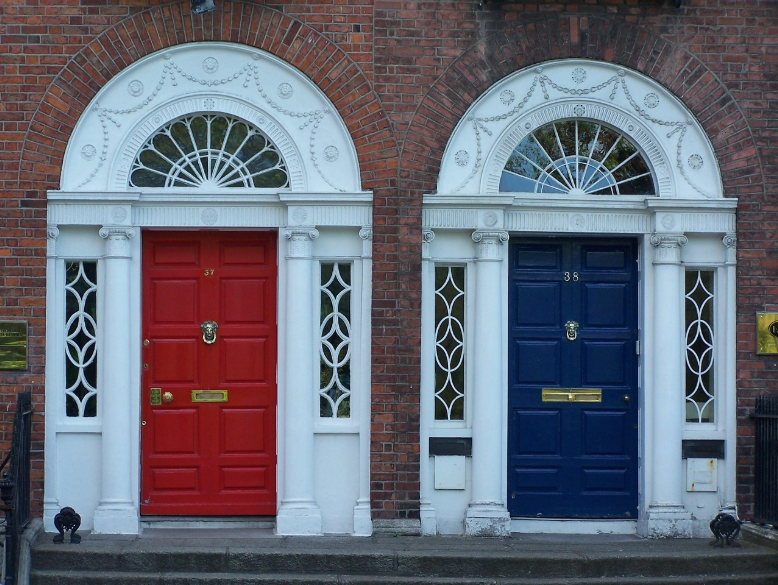 Two adjacent Georgian-style front doors on a brick building, one red (number 37) and one dark blue (number 38), each framed by white columns, ornate fanlight windows, and oval glass panels with white lattice detailing. Both doors feature brass mail slots and knockers, showcasing classic architectural symmetry and contrast in color.