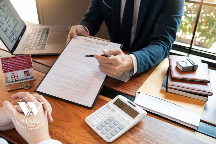 A real estate agent reviews a home loan document with a client at a wooden desk, with a small model house, calculator, and laptop nearby. The Nathan Wenzel Team logo appears in the lower left corner.