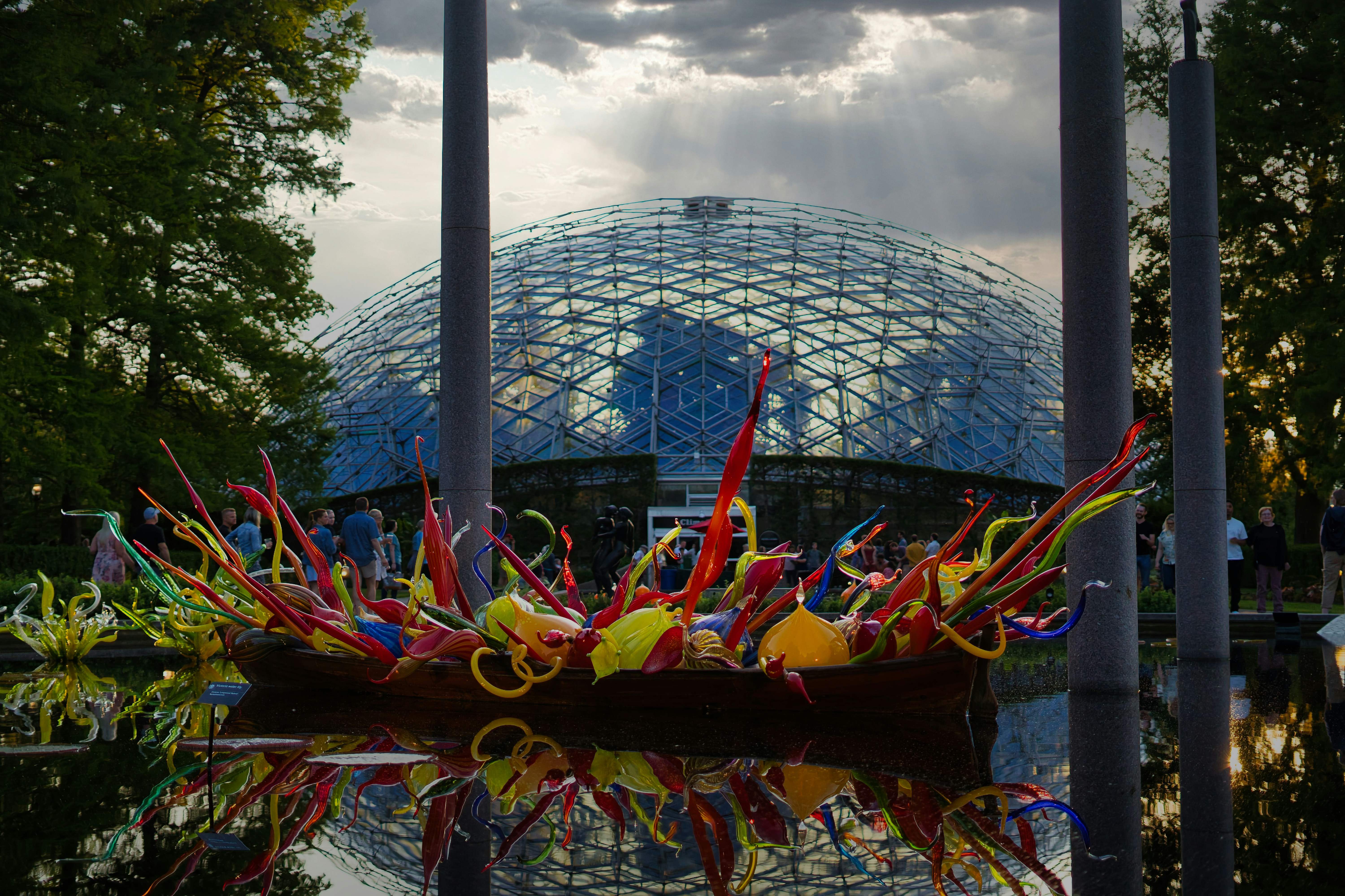 Colorful glass sculpture installation floating on a reflecting pool at the Missouri Botanical Garden in St. Louis, with the iconic Climatron greenhouse dome visible in the background at dusk