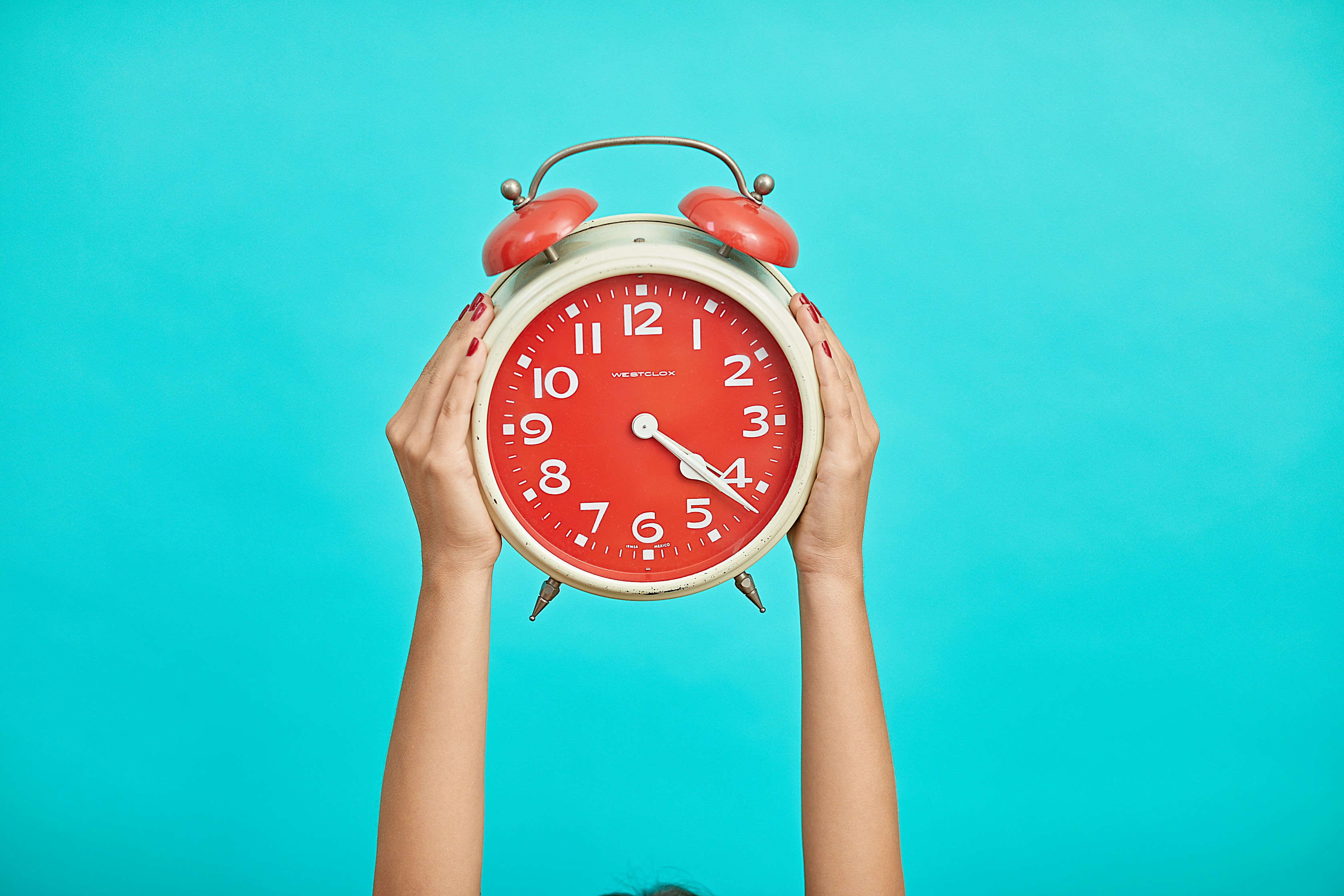 an alarm clock with a red face on a blue background being held up by two hands representing daylight savings time time change