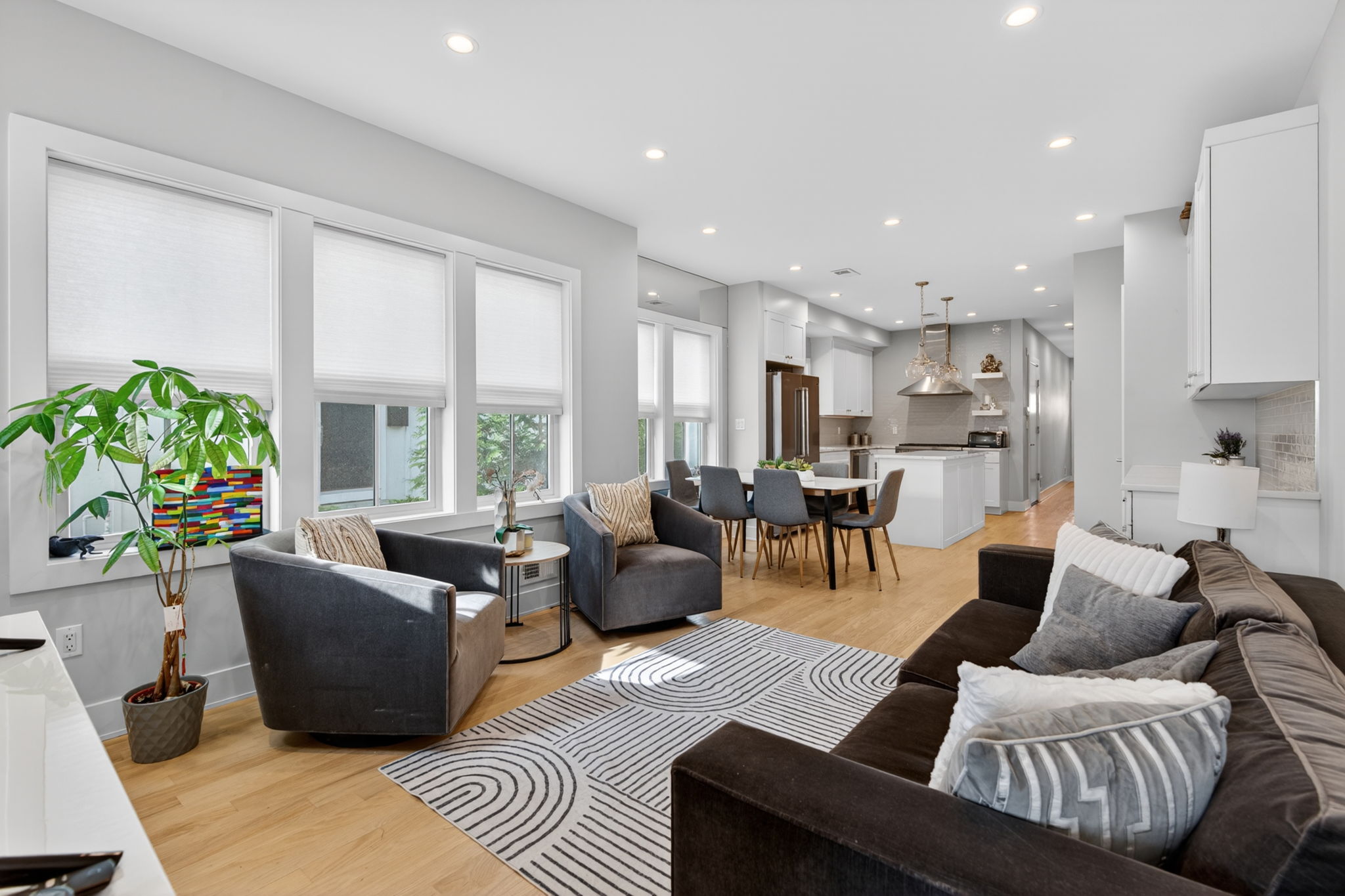 Bright and spacious open-concept living room seamlessly transitions into the dining area and modern kitchen of a newly built 2023 townhouse. The living room features high ceilings, hardwood oak floors, and large windows that flood the space with natural light. The dining area showcases elegant baseboard molding and custom built-in storage with quartz countertops. The crisp white kitchen is equipped with recessed lighting, stainless steel appliances, quartz countertops, and custom cabinetry, creating a stylish and functional heart of the home with 4 bedrooms, 4 bathrooms, plus a half bathroom..