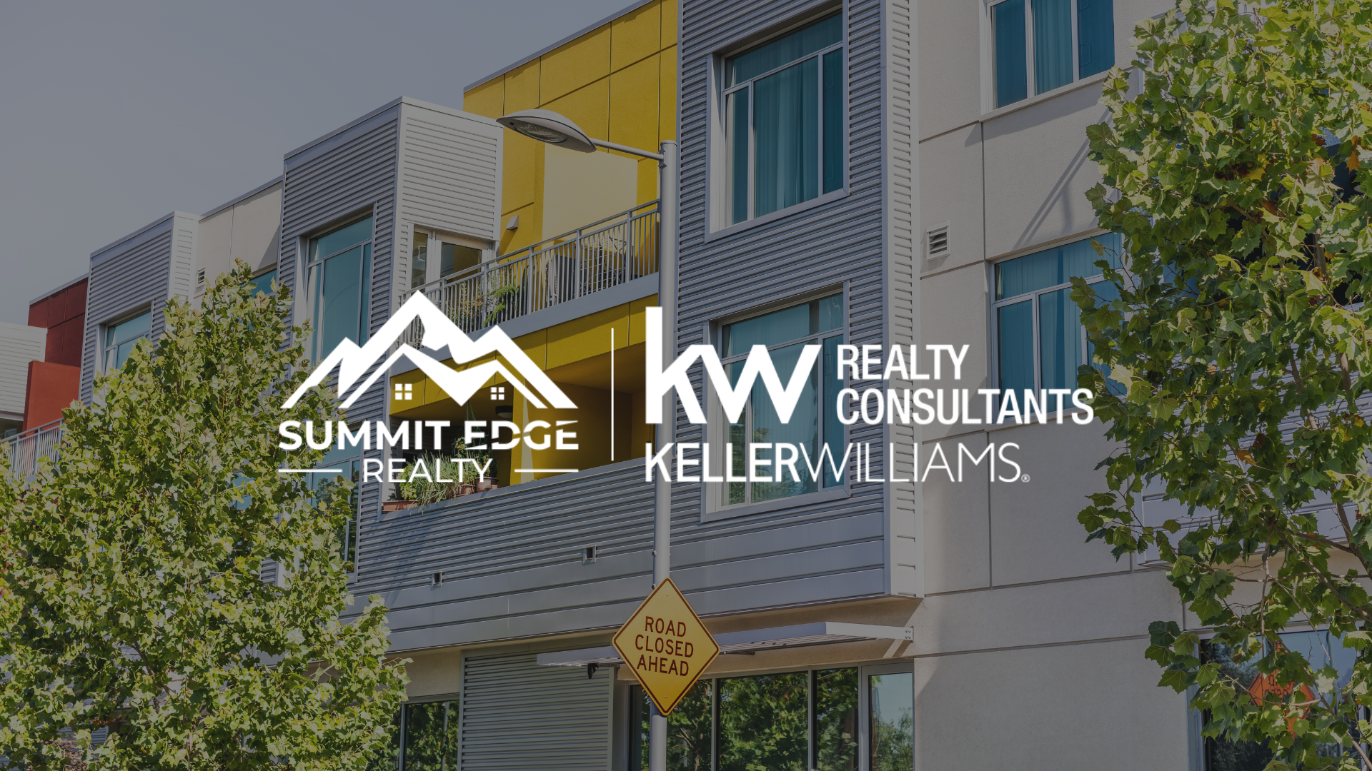 Exterior view of a Harrison County, IN, multifamily housing community featuring beige and brown townhouses, each with an attached garage and a small front yard with greenery, along a peaceful street, branded with the Summit Edge Realty and KW Realty Consultants Keller Williams logo in white.