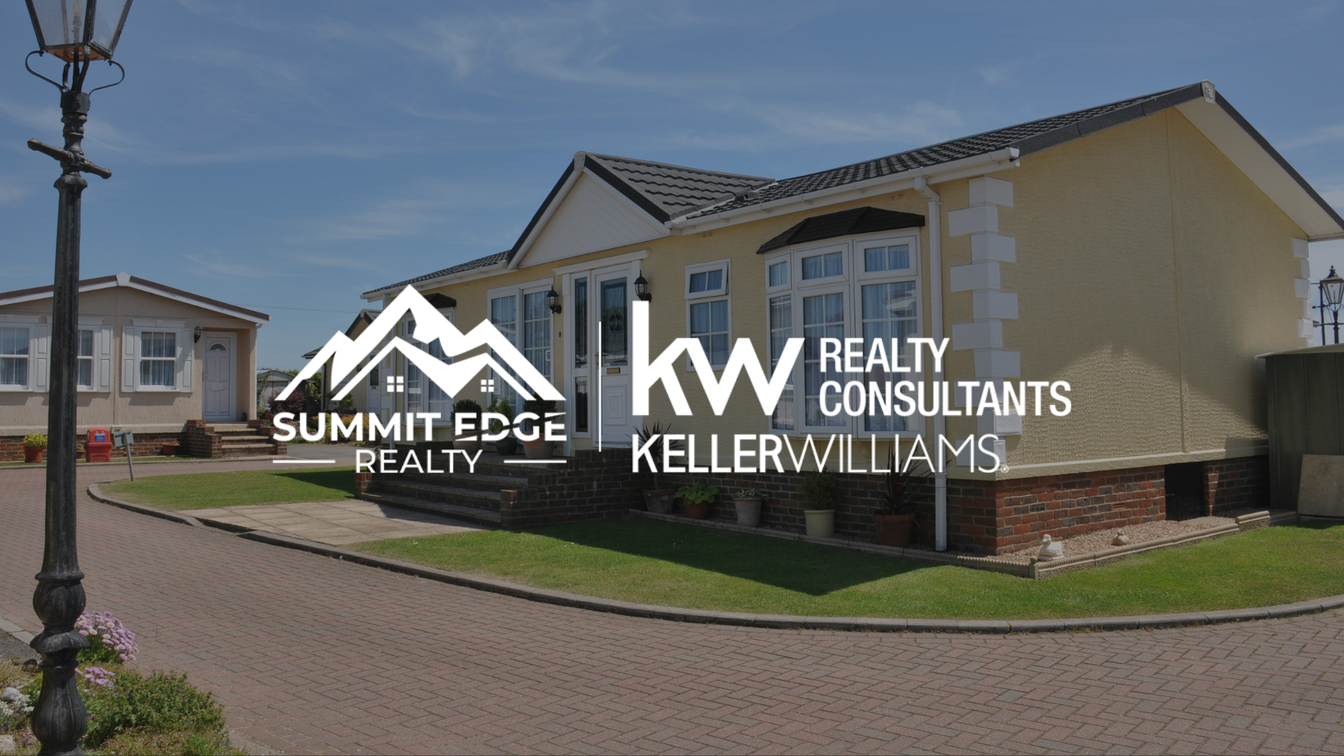 Image of a manufactured home in Louisville, KY, highlighting a beige exterior with prominent bay windows, a brick base, and a paved walkway with a lush green lawn, under a clear blue sky, displaying the Summit Edge Realty and KW Realty Consultants Keller Williams logo in white text.