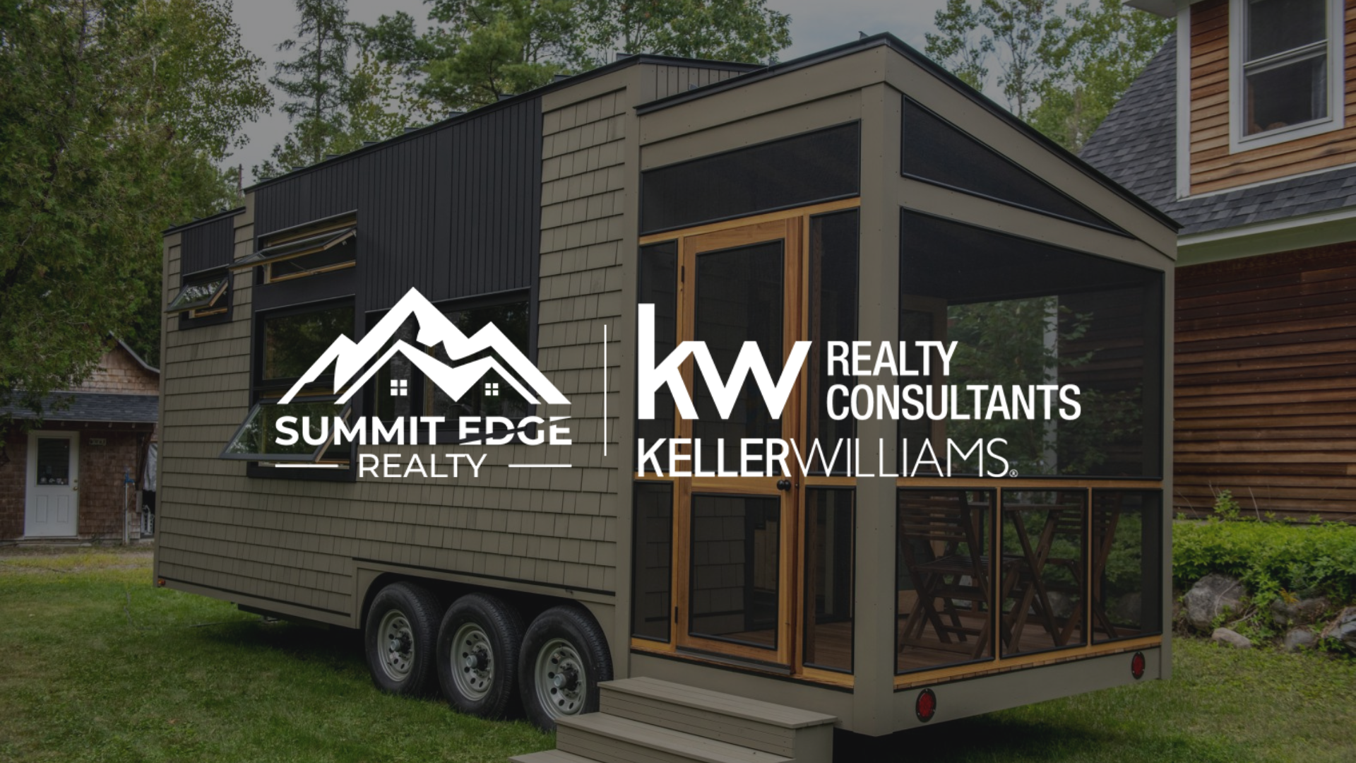 Exterior view of a Harrison County, Indiana, tiny house featuring a gray wooden facade, a sloped green roof, and a small deck with railings, situated on a dirt site with trees and shrubs under a clear sky, branded with the Summit Edge Realty and KW Realty Consultants Keller Williams logo in white.