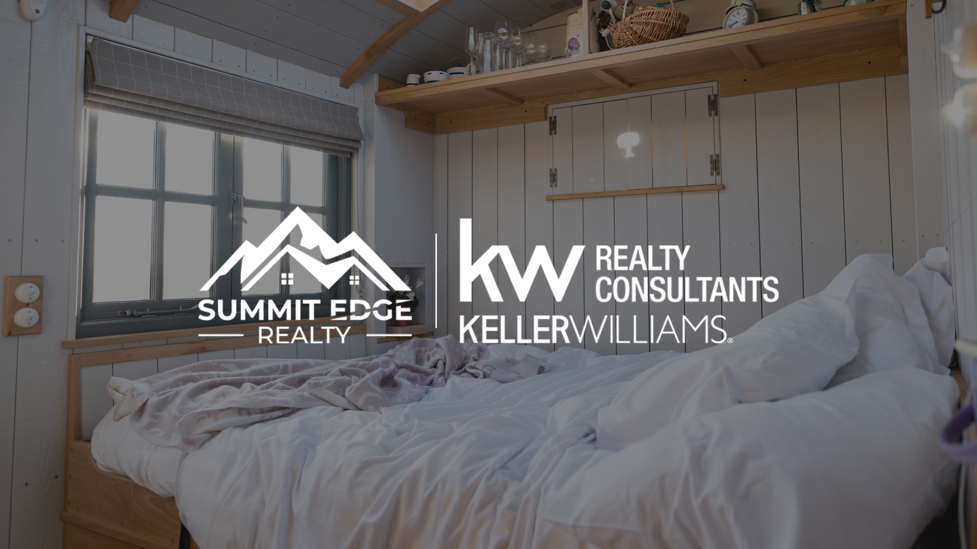Interior view of a cozy tiny home featuring a neatly made bed with white and gray bedding, a wooden shelf with decorative items, and large windows with blinds, set against light wood paneling, overlaid with the Summit Edge Realty and KW Realty Consultants Keller Williams logo in white text.